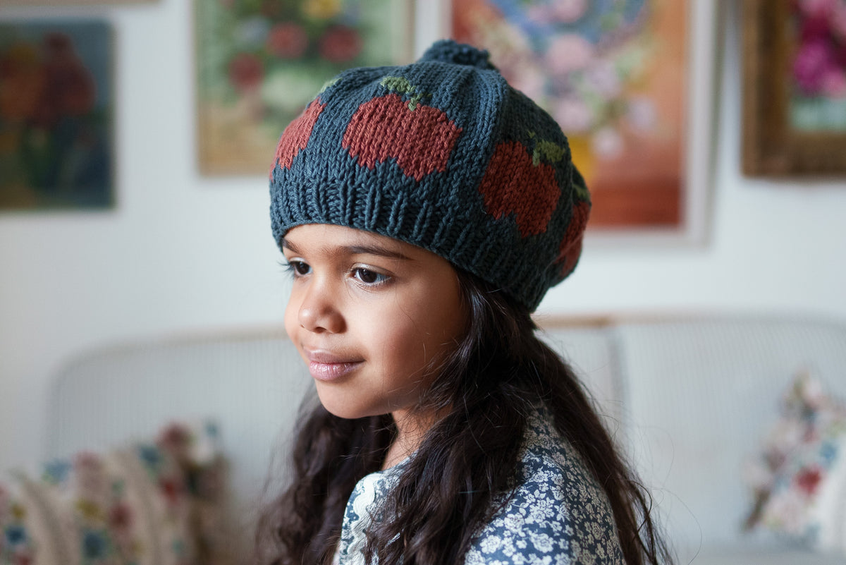 Young girl wearing hand-knitted beret  with bobble, with charming red apple pattern, against a sofa with floral cushions and vintage paintings, in a natural children's clothing setting.