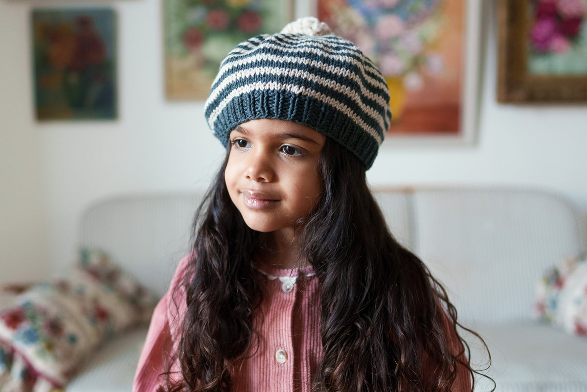 Young girl wearing pretty cord vintage-inspired dress and hand-knit black and cream striped beret in front of a sofa and floral paintings, embodying natural and ethical children's clothing.