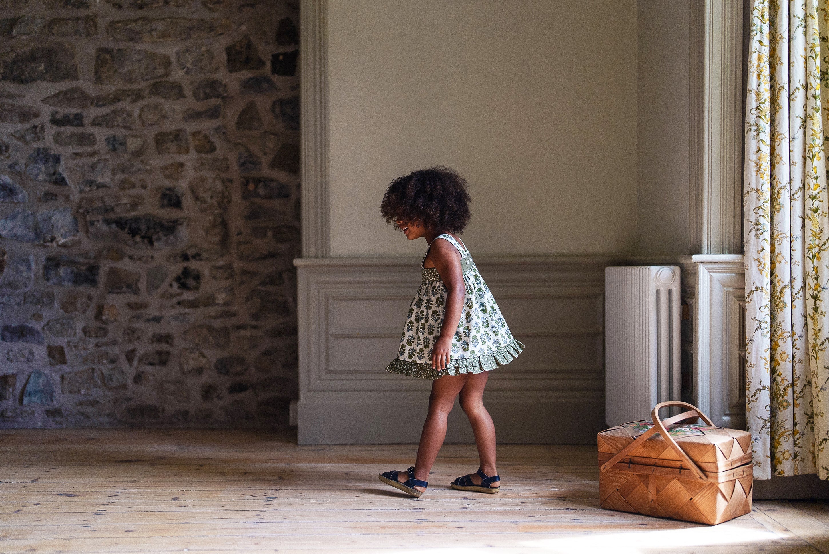 Young child wearing a vintage style organic cotton sun top with a green floral print, walking barefoot on wooden floor in a cozy, natural light room with stone wall and wicker basket.