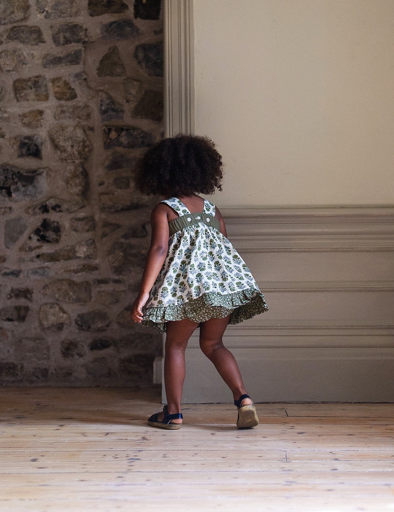 Toddler wearing a vintage style sun top in organic cotton with a green floral print, walking on a wooden floor by a stone and plaster wall in a softly lit room.