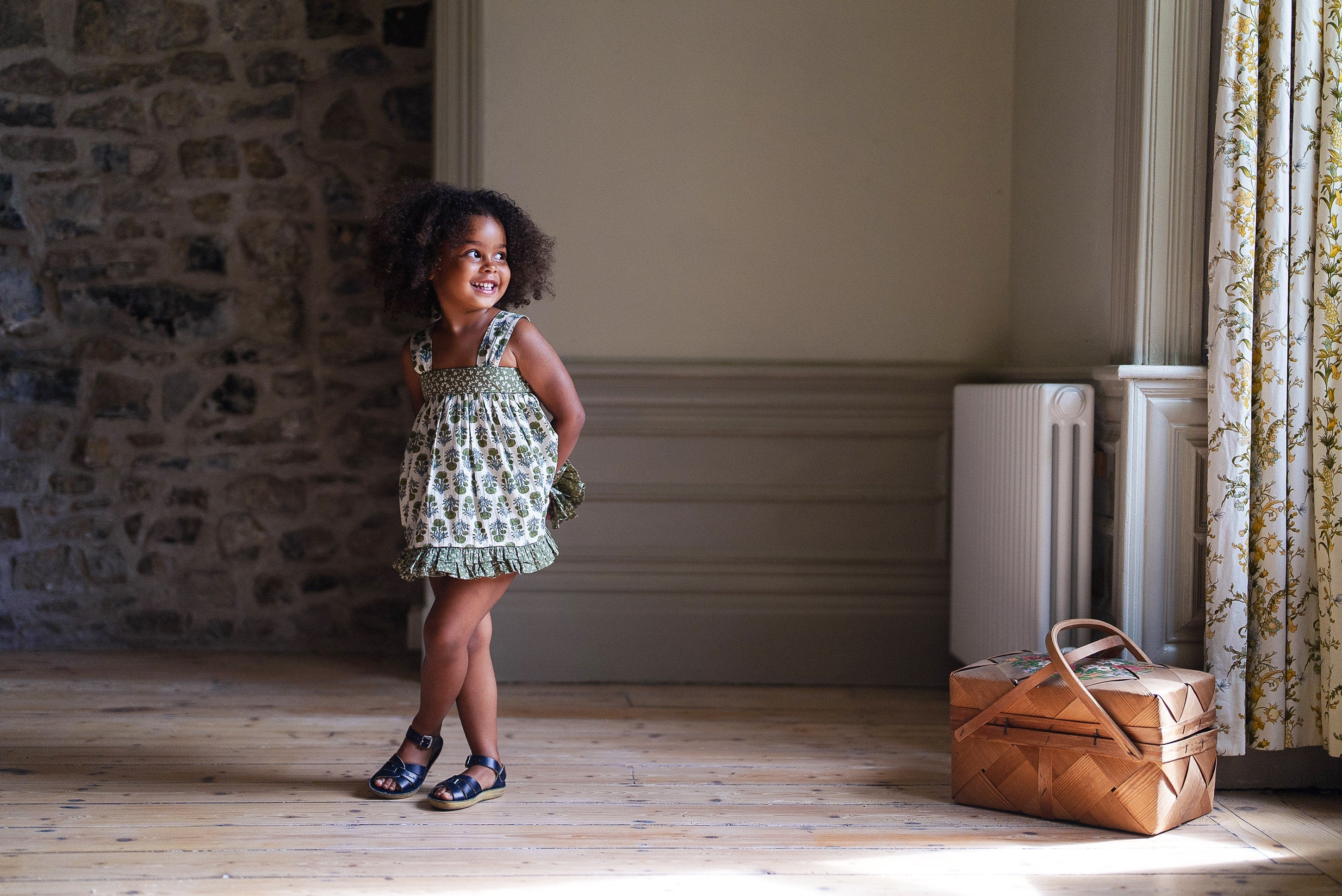 Young girl wearing a vintage style organic cotton sun top in green floral print standing in a rustic room with natural light, showcasing sustainable, ethically made children's clothing.