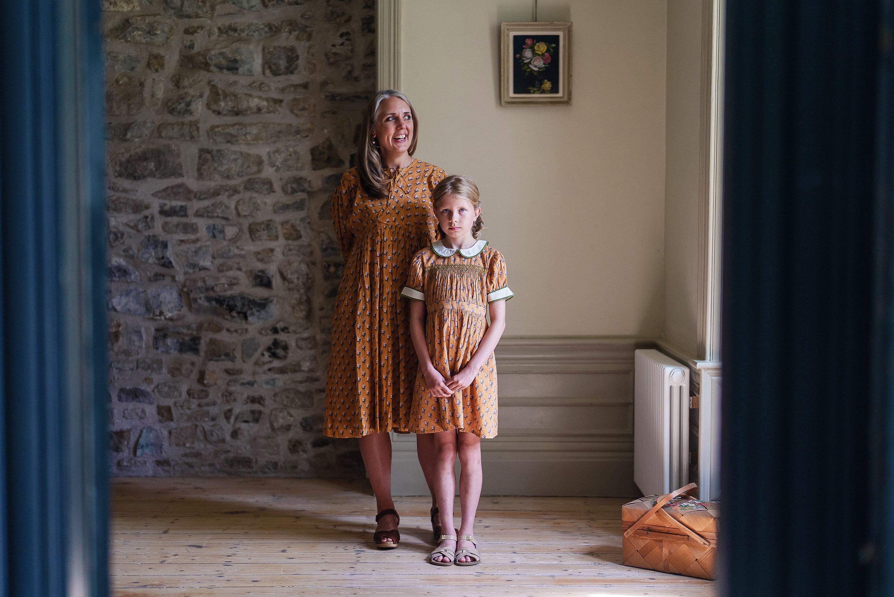 A woman and a young girl wearing ethically made vintage style dresses in warm saffron floral patterns, standing in a softly lit room with natural wooden floors, reflecting slow, natural childhood fashion.