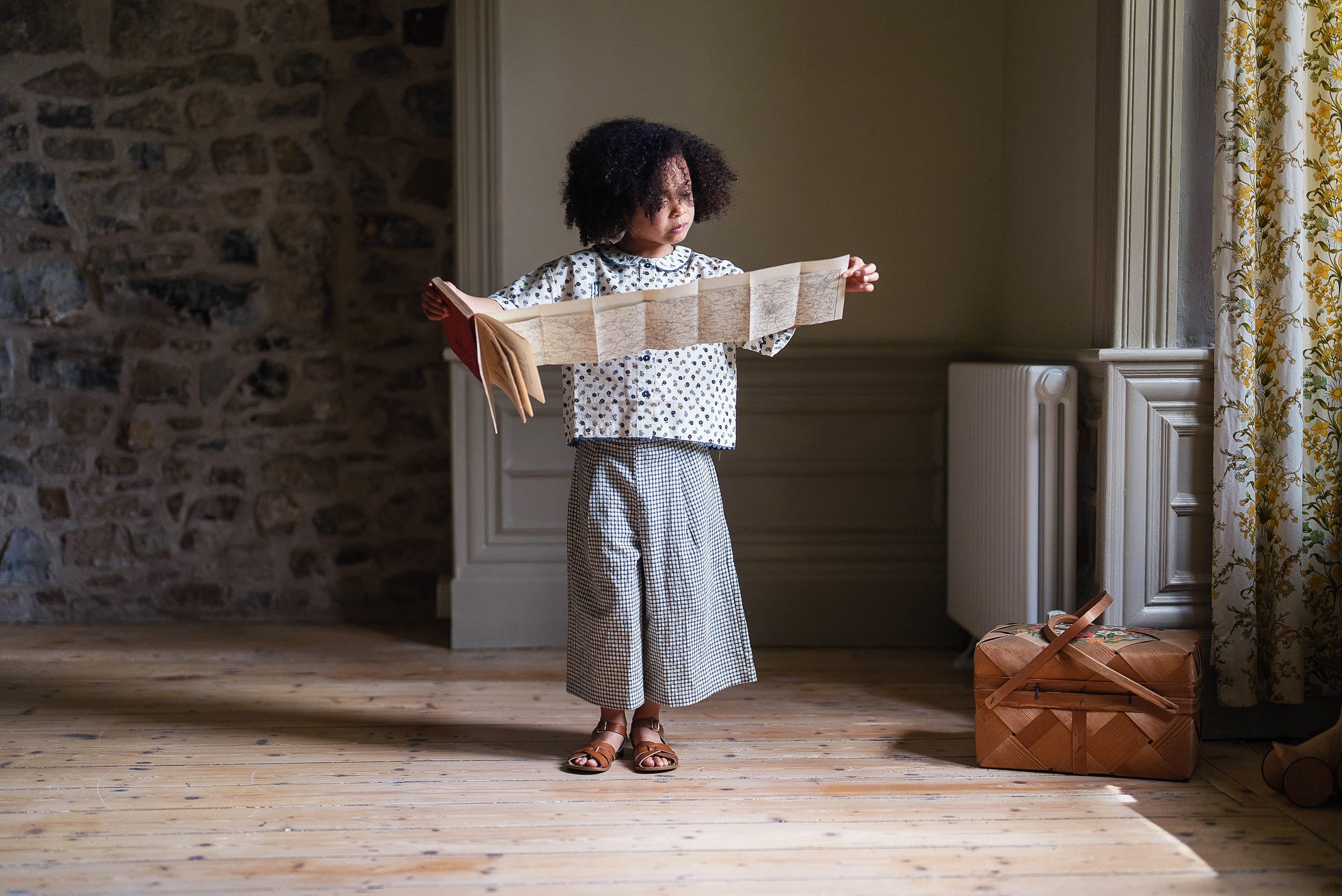 Child wearing a vintage style floral cotton blouse and wide-legged trousers standing indoors holding an unfolded map, bathed in natural light by a window with floral curtains.