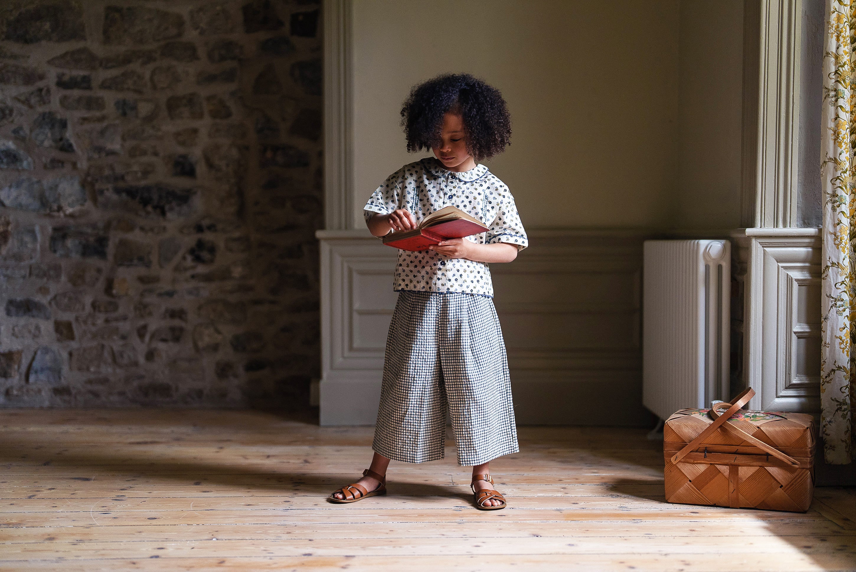Child wearing a vintage style girls blouse and wide-leg cotton pants, standing indoors with natural light from a window, showcasing sustainable and organic children’s clothing.