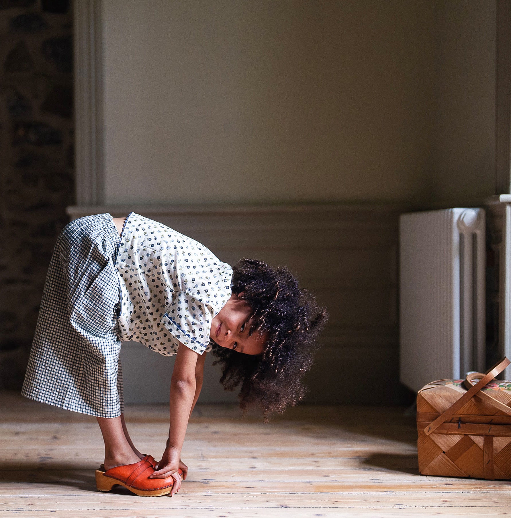 Child wearing organic cotton daisy floral blouse and gingham skirt, bending forward indoors near wooden floor and woven basket, showcasing vintage style children's clothing.