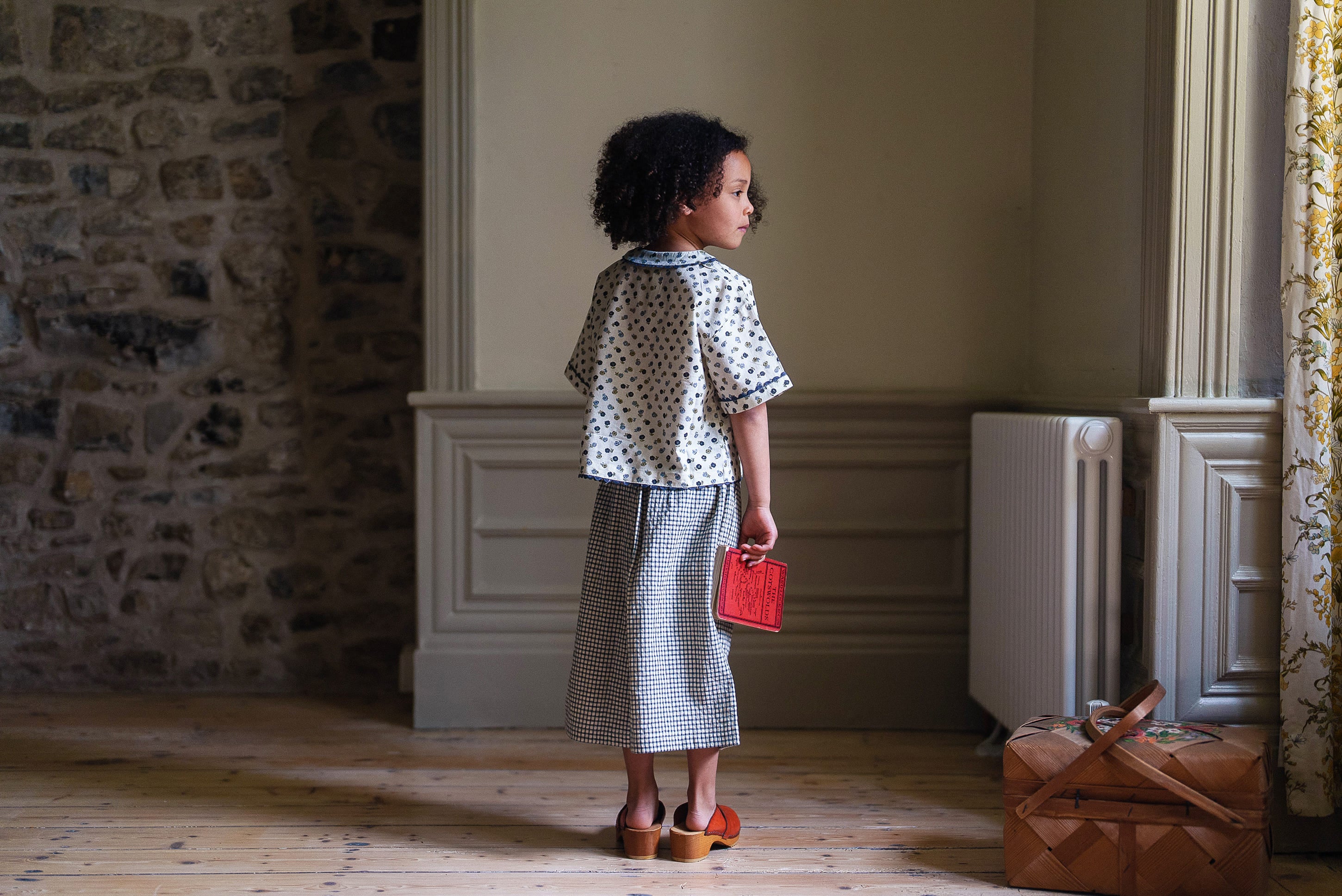Child wearing a cotton blouse with a delicate floral pattern and a long skirt, standing in a sunlit vintage-style room holding a small red purse, showcasing ethical, organic children’s clothing.