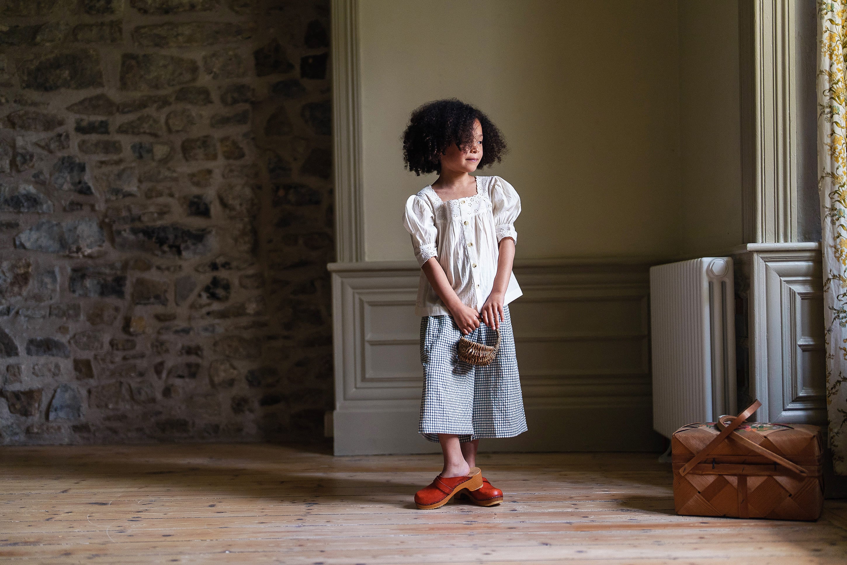 Young girl wearing an off-white cotton vintage style blouse and skirt, standing in a softly lit room with natural wooden floors and stone walls, holding a small basket.