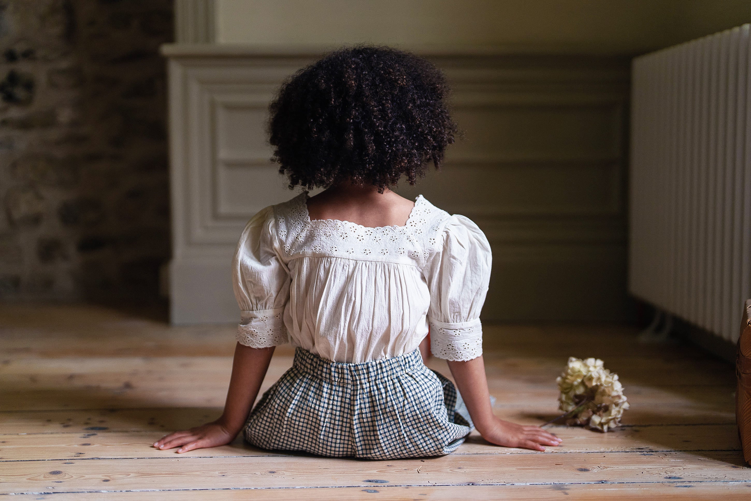 Child wearing an off-white cotton vintage-style blouse with puffed sleeves and lace details, sitting on a wooden floor in a softly lit, natural setting.