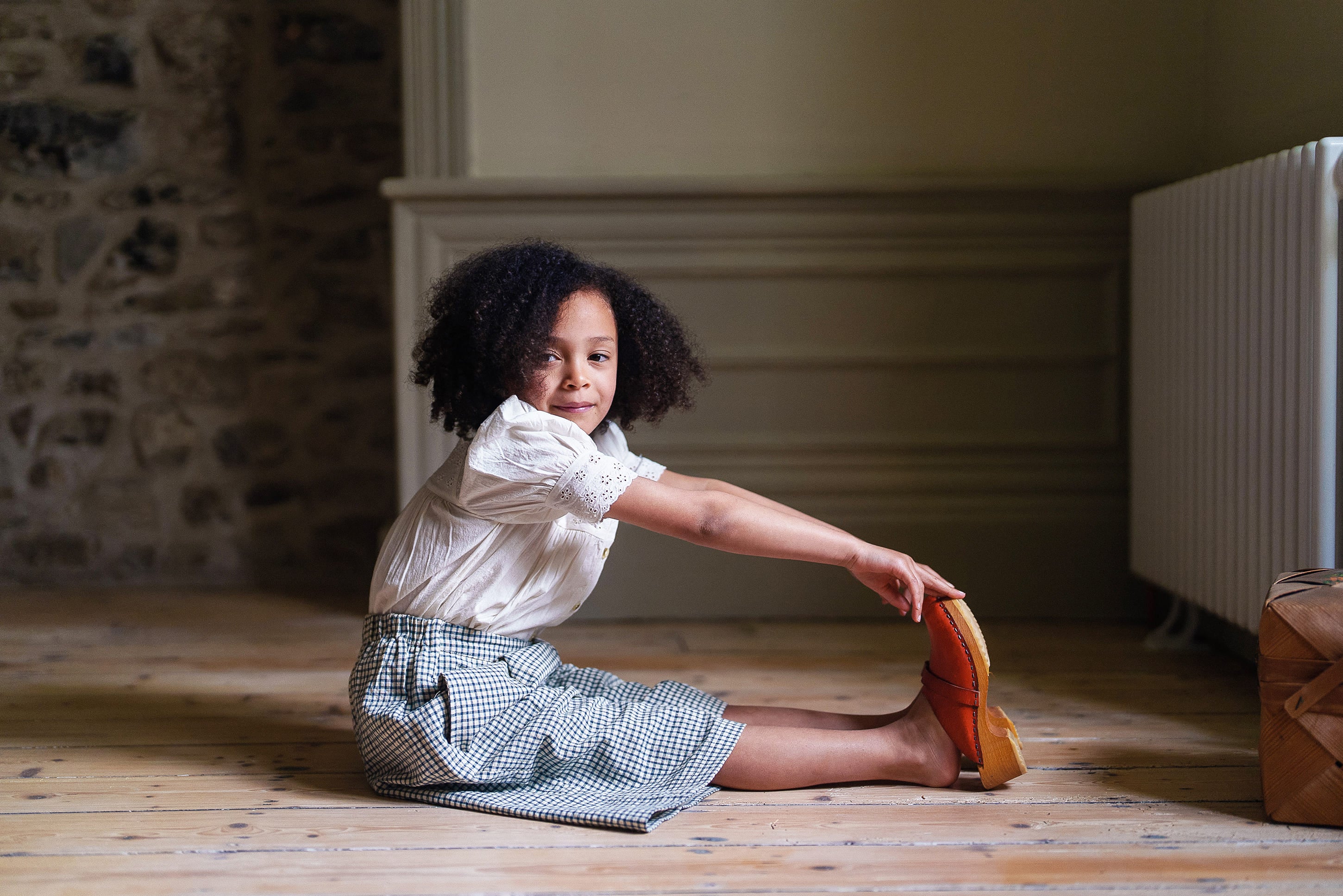 Young girl stretching indoors wearing a vintage style cotton blouse and checked skirt in a natural, ethically made children's clothing setting