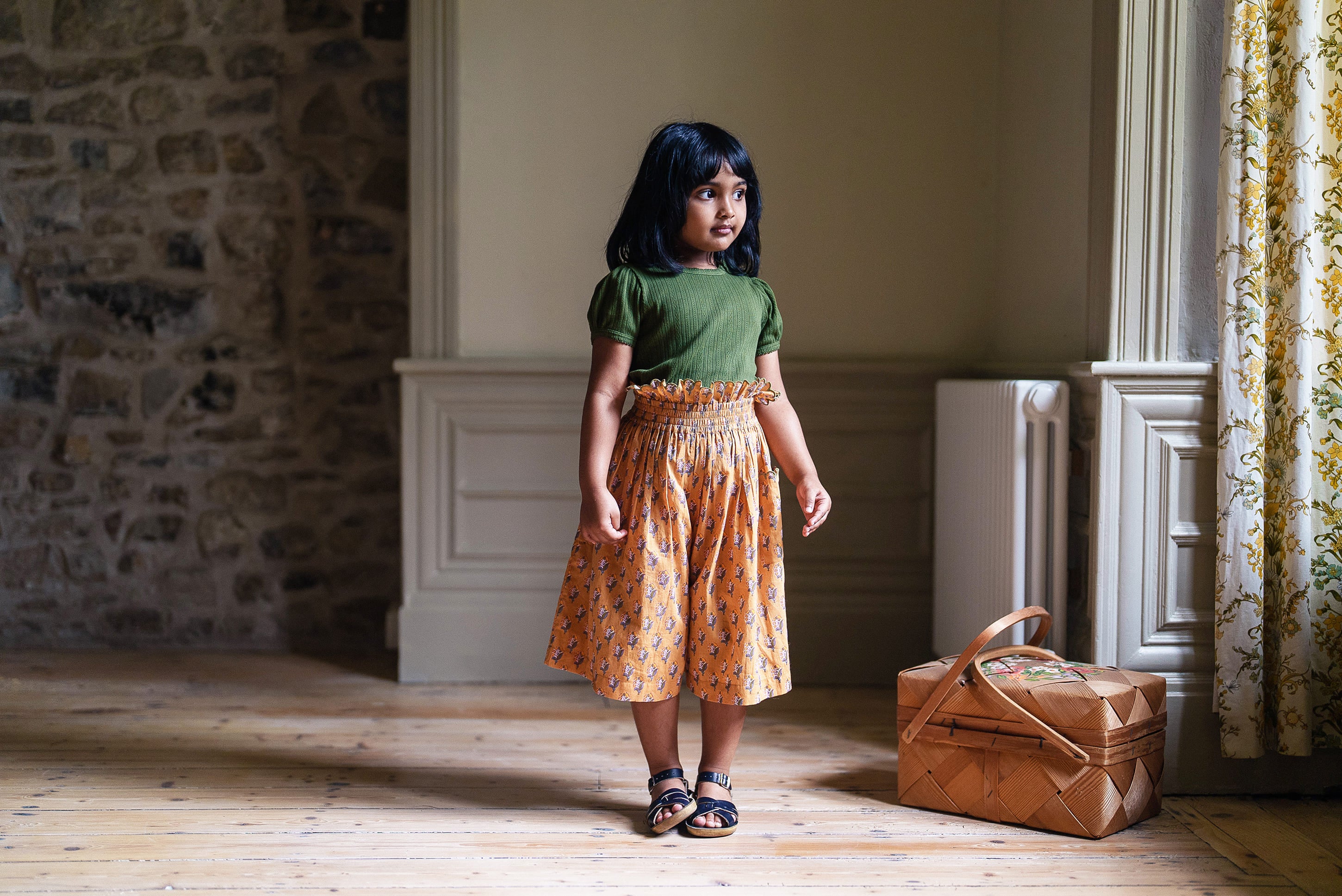 Young girl wearing a vintage-style organic cotton green top and patterned skirt, standing in a naturally lit room with wooden floors and a woven basket nearby.