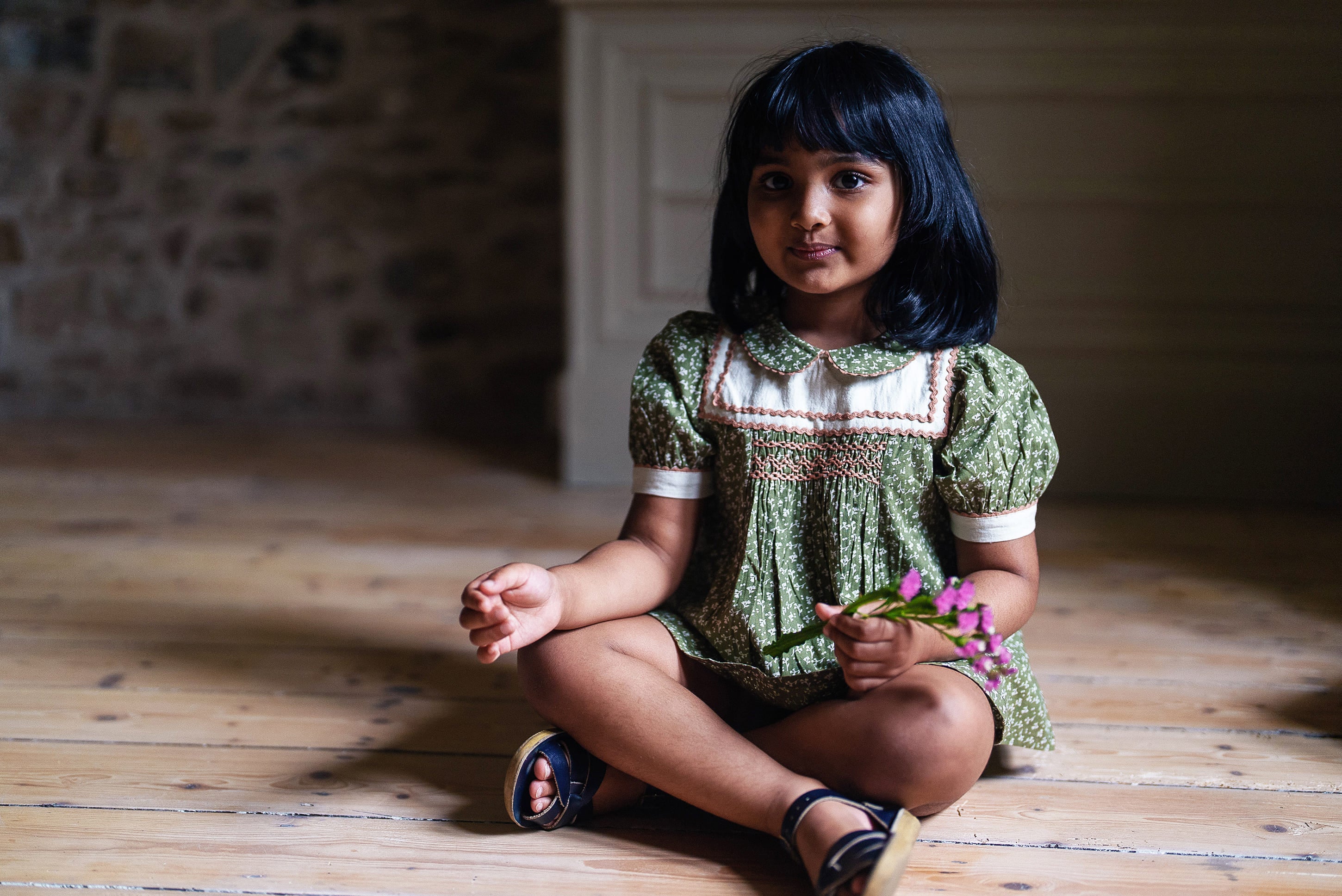 Young girl wearing a vintage style green cotton blouse with smocked detailing, sitting on a wooden floor holding small purple flowers, showcasing sustainable children's clothing.