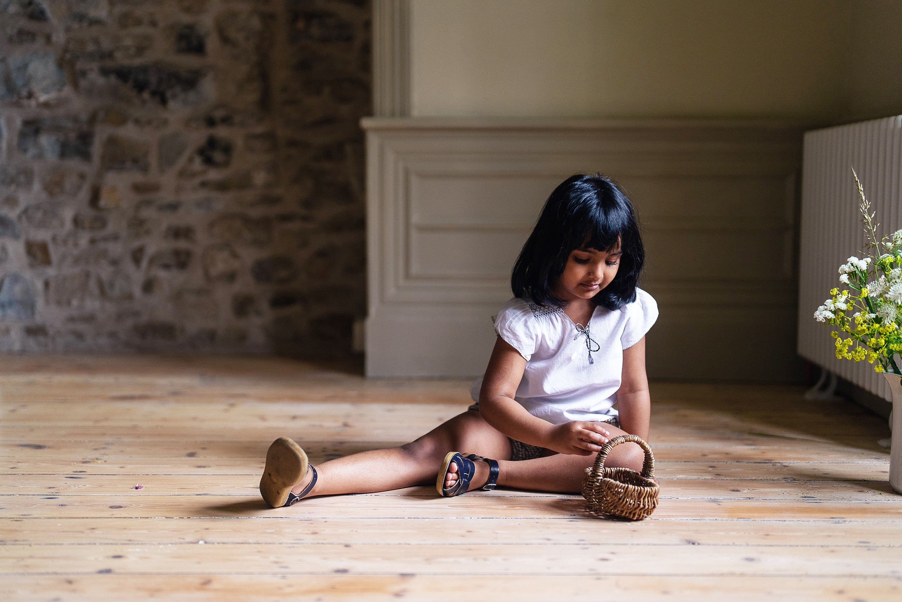 Young girl wearing an off-white vintage style blouse sitting on a wooden floor, playing with a small wicker basket in a naturally lit room with rustic stone walls.
