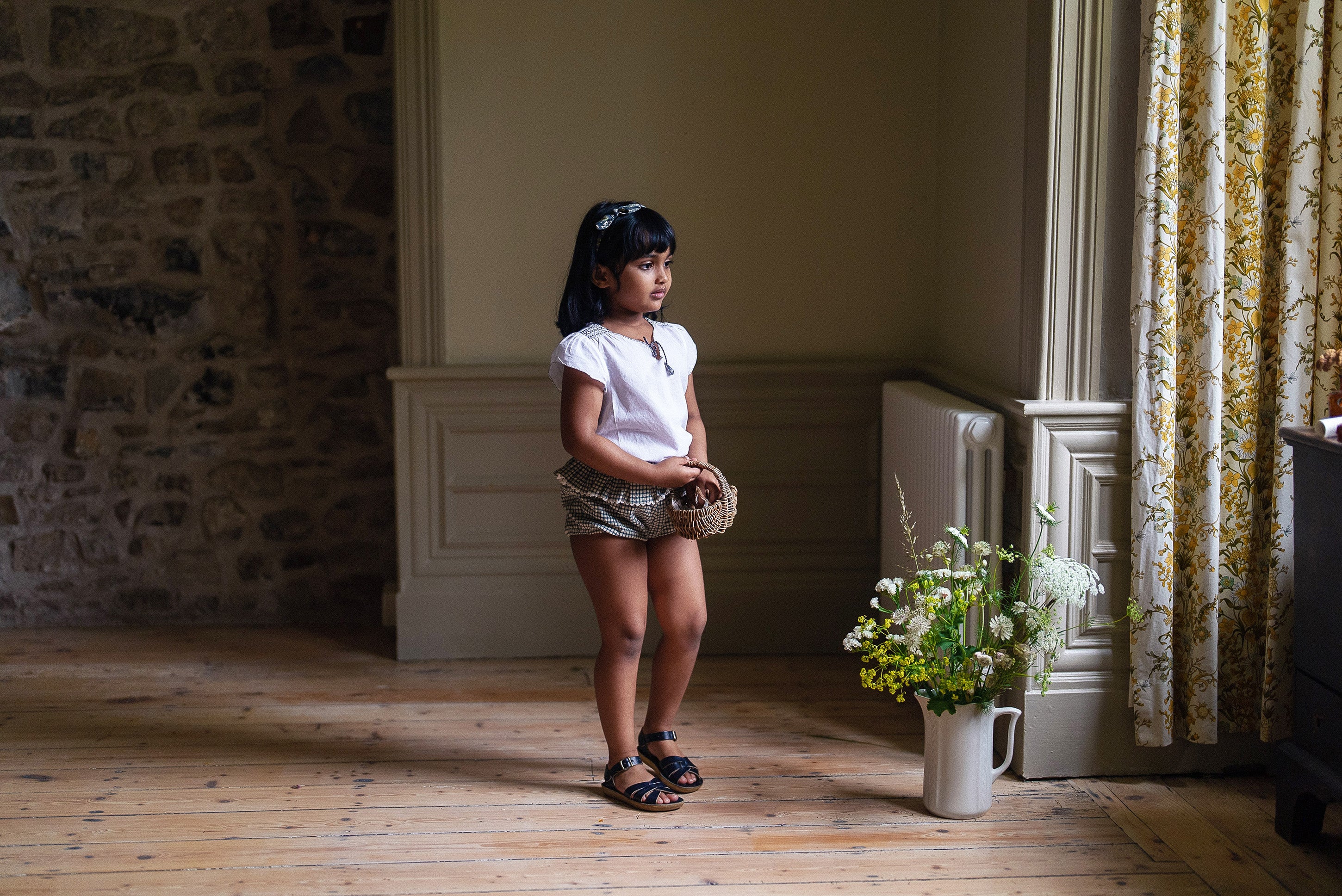 Young girl wearing a natural off white vintage style blouse paired with shorts, standing in a softly lit room with wooden floors and a floral arrangement by the window.