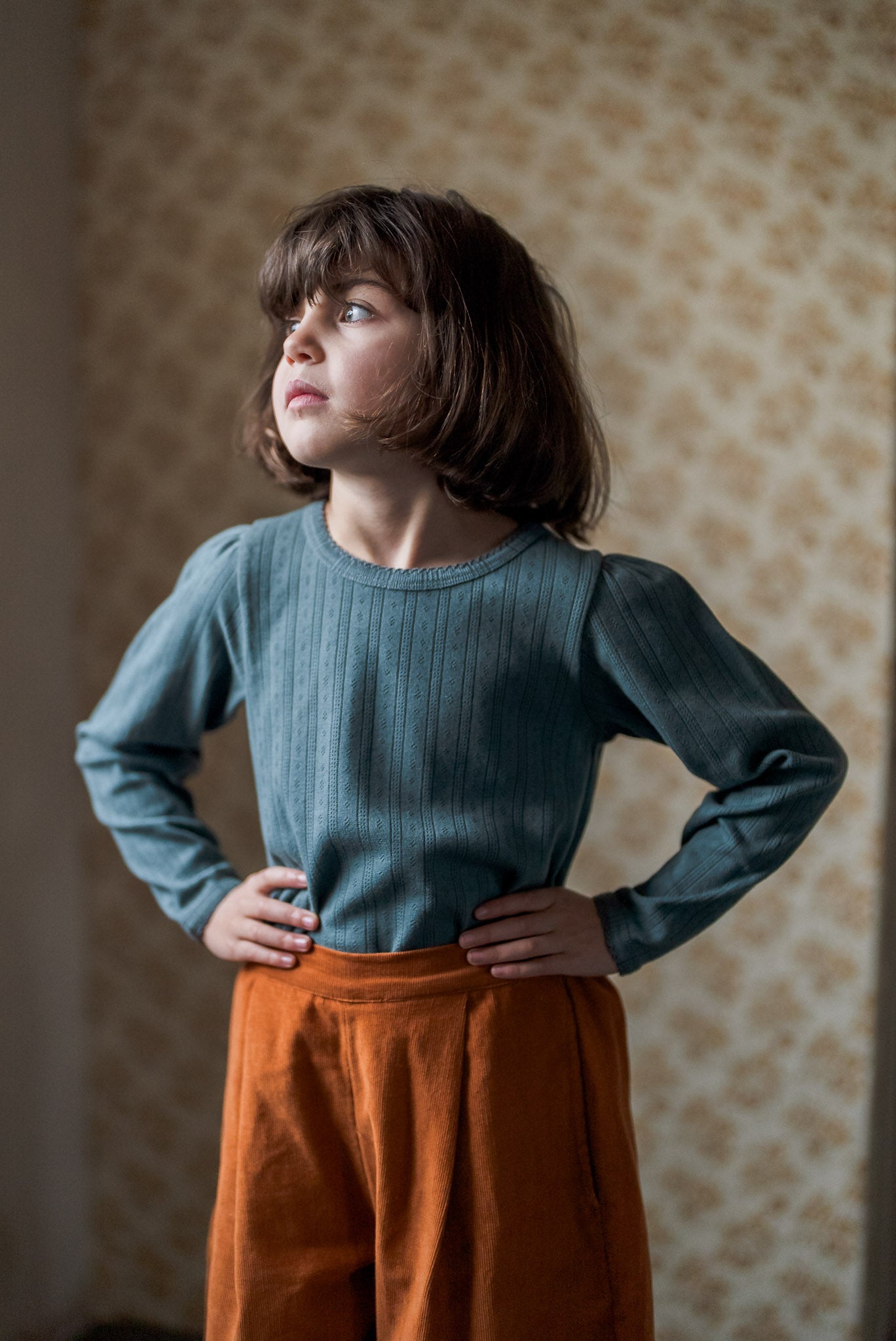 Young child wearing a soft navy blue cotton t-shirt with subtle ribbed texture and vintage-style burnt orange pants, standing thoughtfully in a warm, natural light setting.
