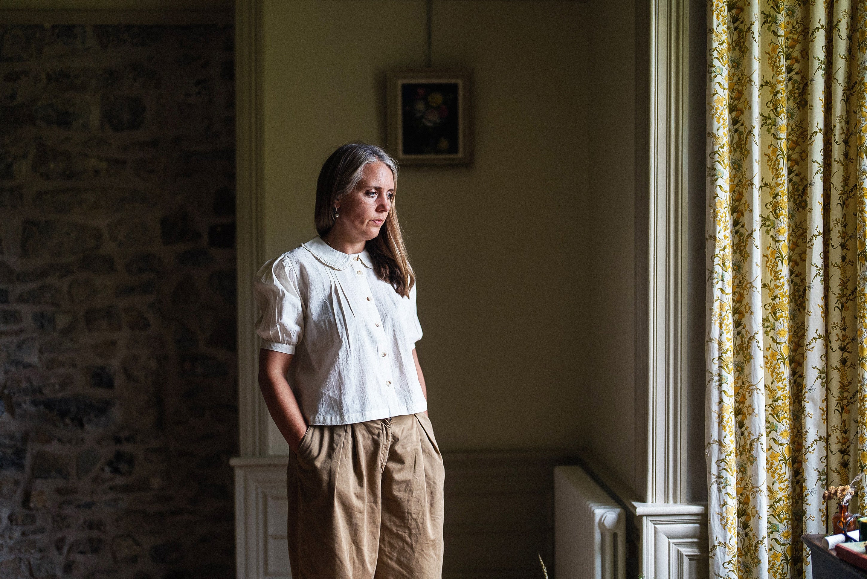 Woman wearing a white cotton blouse with vintage-inspired design details, standing thoughtfully by a window in a softly lit, cozy room with natural materials and warm tones.