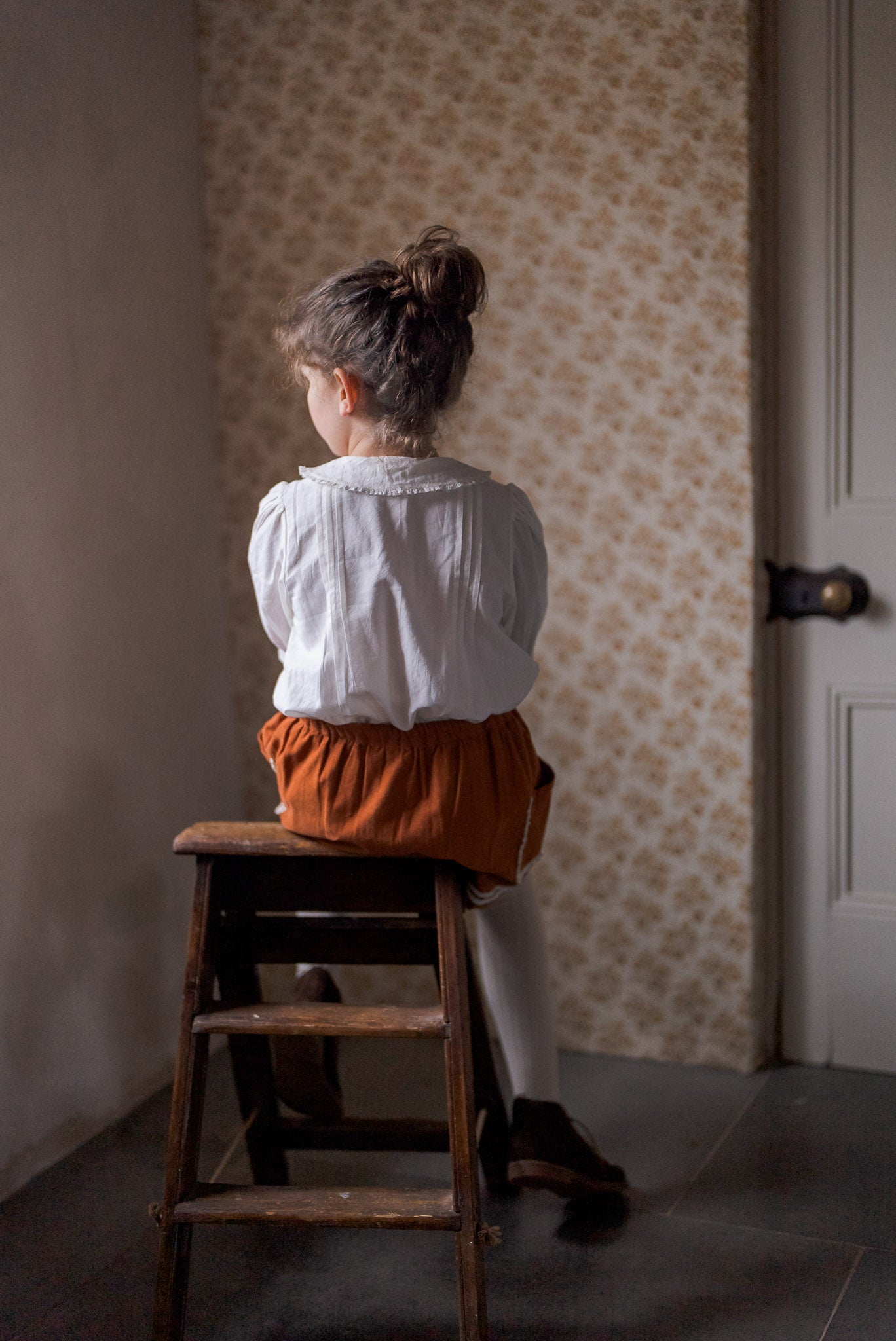 Young child wearing an off-white cotton blouse with vintage style details, sitting on a wooden stool in a softly lit, traditionally styled room, embodying sustainable kids clothing.