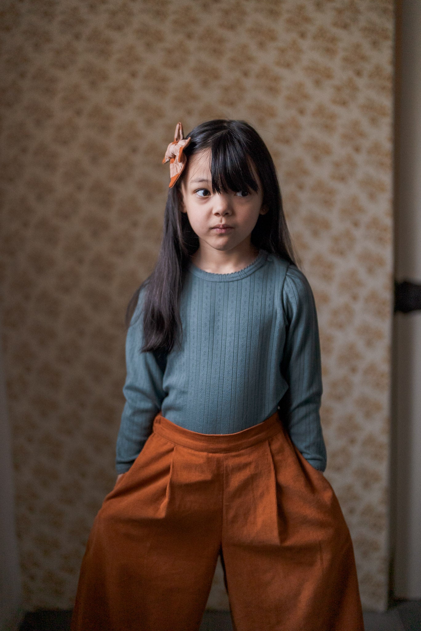 Young girl wearing a vintage-style navy blue cotton T-shirt and rust-colored wide-leg pants, styled with a natural fabric bow in her hair, standing indoors.