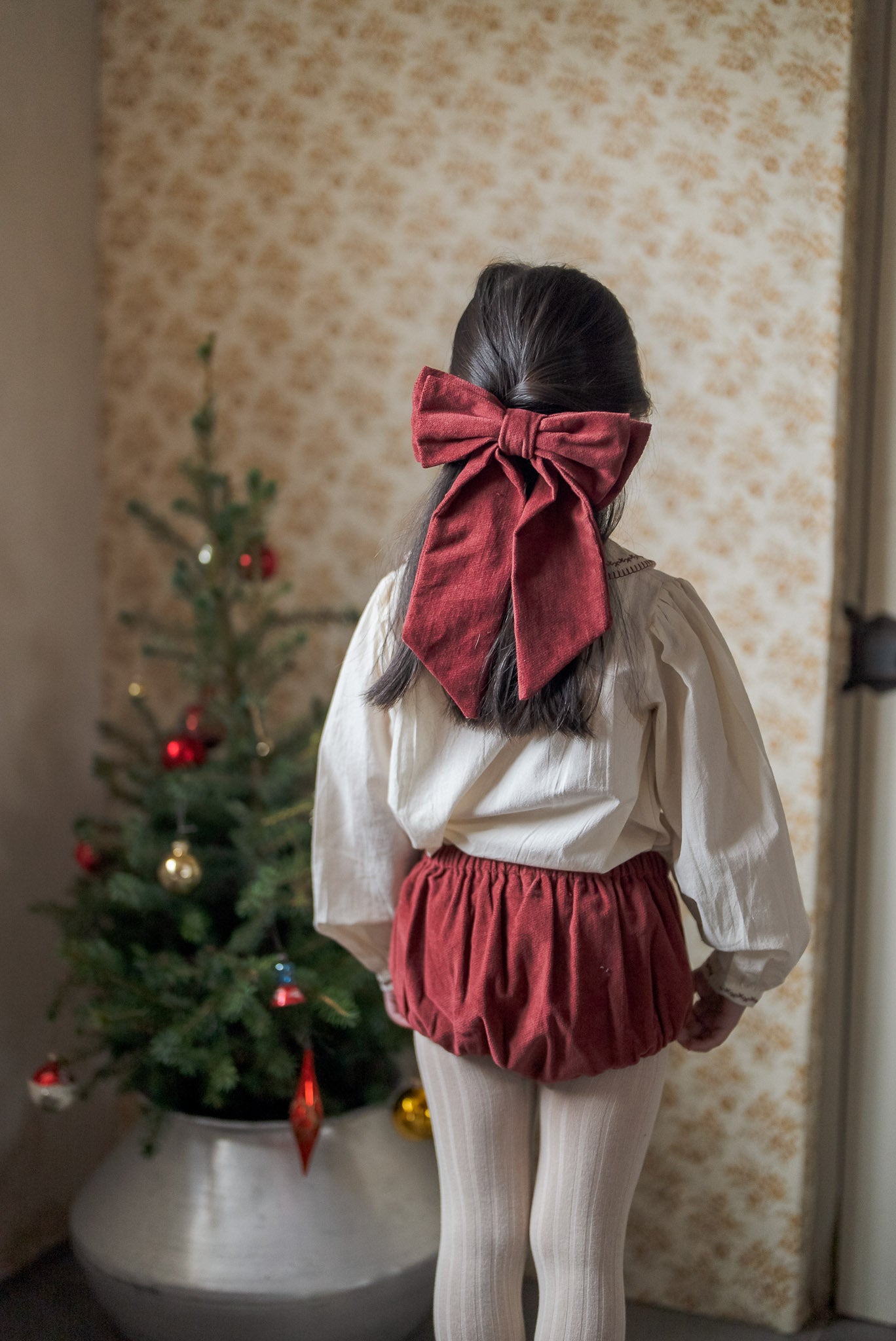 Young girl wearing a vintage style red velvet bow clip in her hair, paired with a classic cream blouse and red bloomers, standing by a decorated Christmas tree.