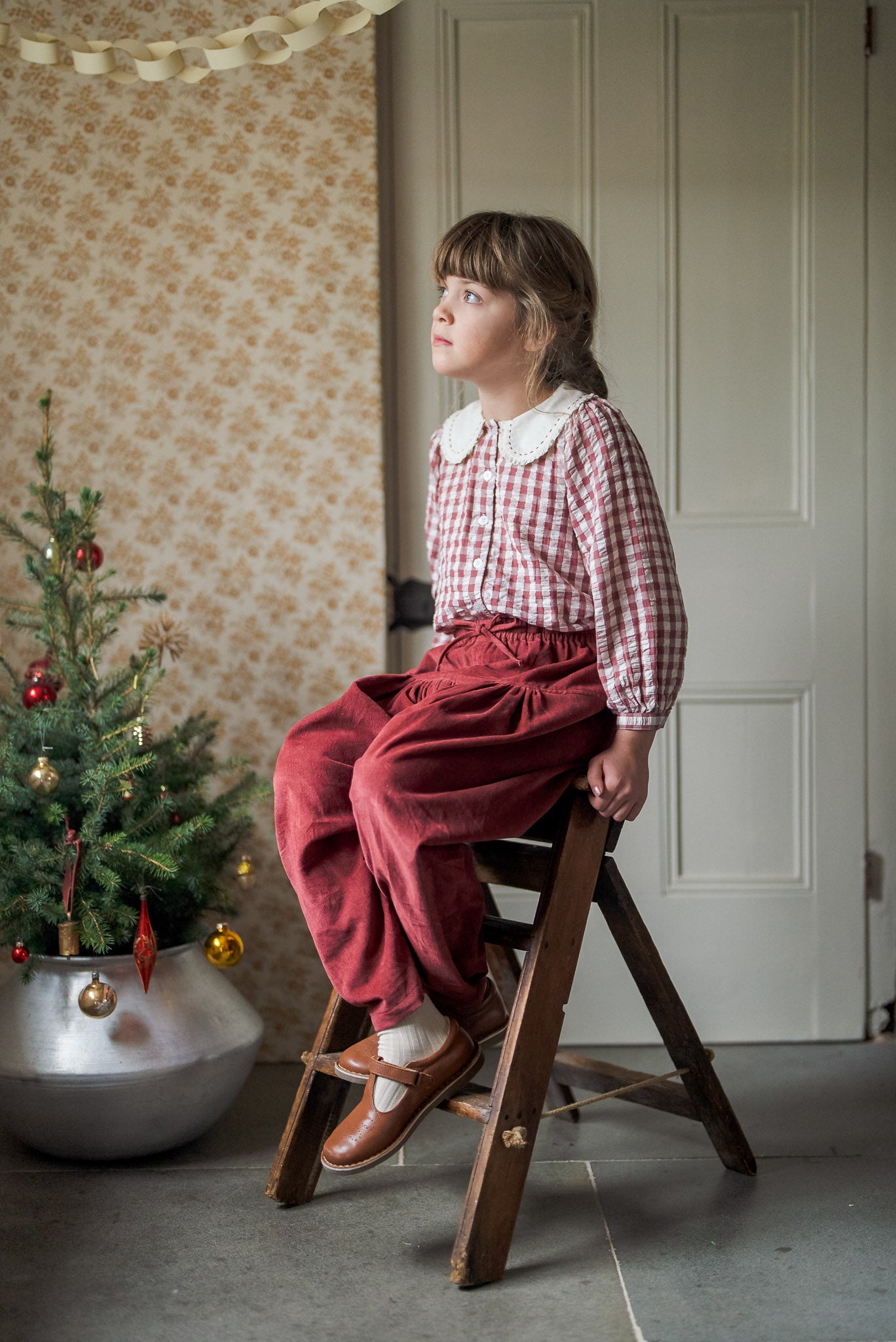 Young girl wearing a vintage-style red and white checkered cotton blouse with a lace collar and rust-colored pants, sitting on a wooden step ladder near a small decorated Christmas tree in a cozy room with floral wallpaper.
