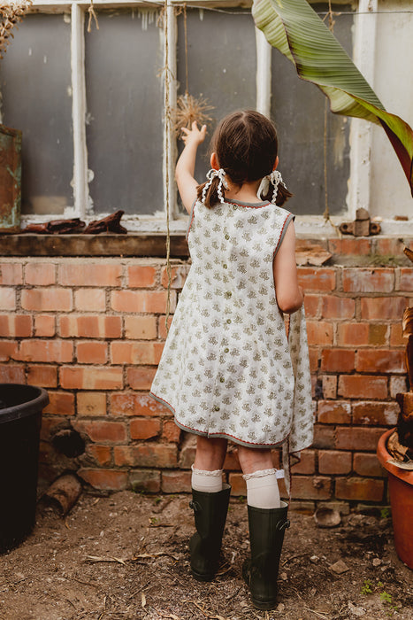 Model wears A-line girl's dress in organic, white floral cotton fabric. The back of the dress has buttons and green binding with pink ric-rac trim. Model wears green  wellie boots and frilly knee high socks.