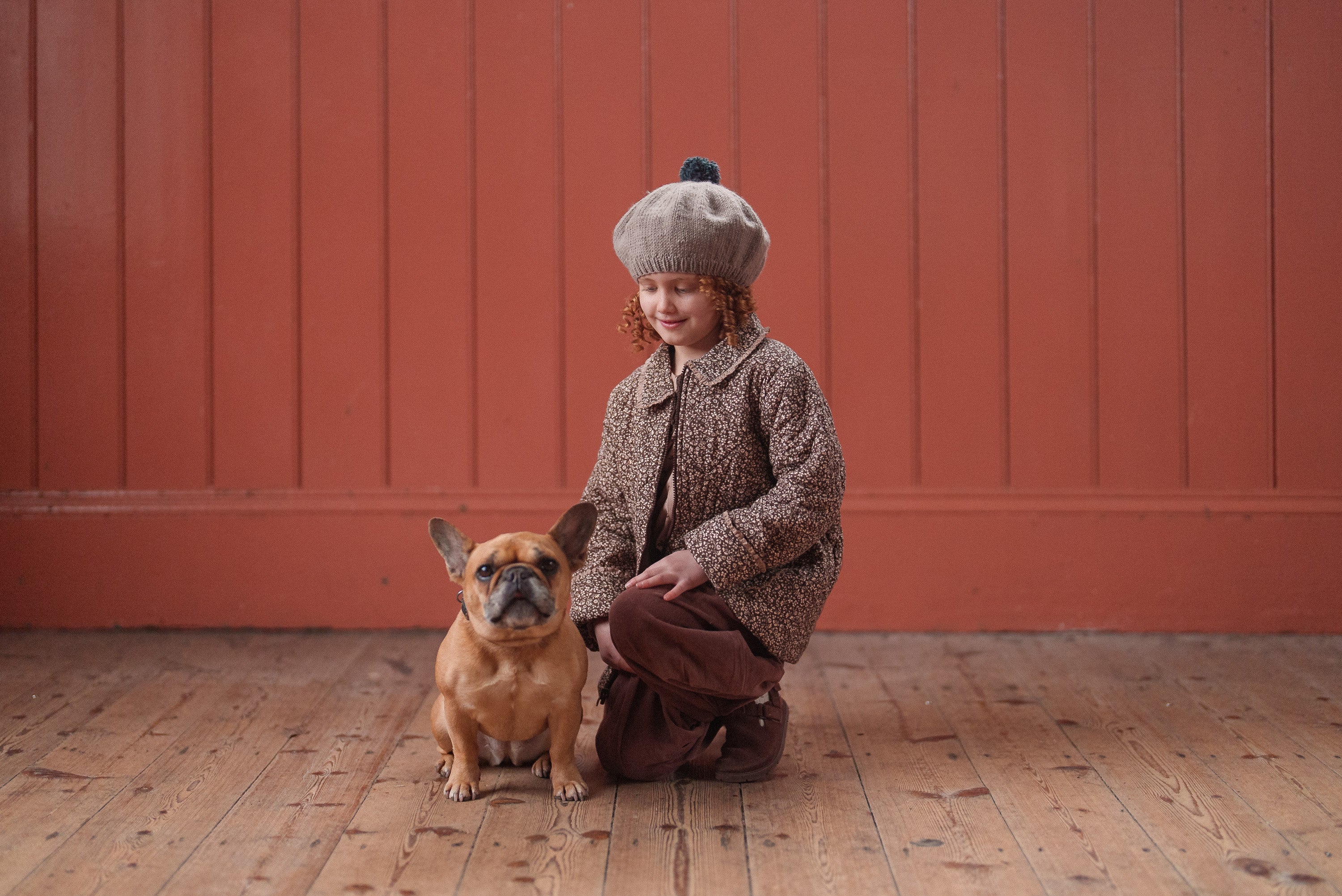 Young child in vintage-style floral brown jacket and beige beret crouches beside a small French bulldog on wooden floor against reddish wall, wearing sustainable organic cotton kids clothing.