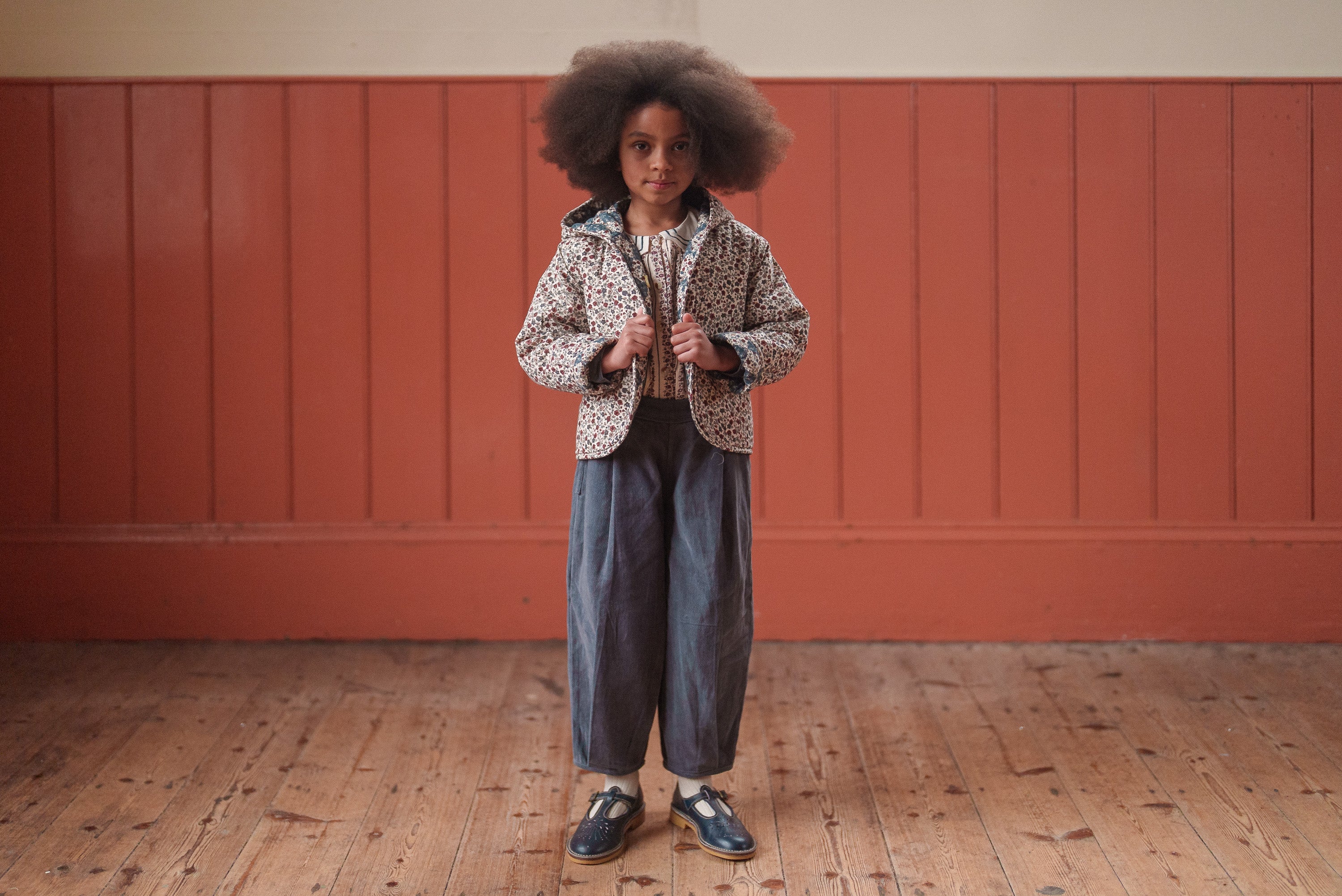 Child wearing vintage-style children's clothing featuring blue velvet trousers made from organic cotton, standing on wooden floor against a warm-toned wall in an ethically made, sustainable outfit.