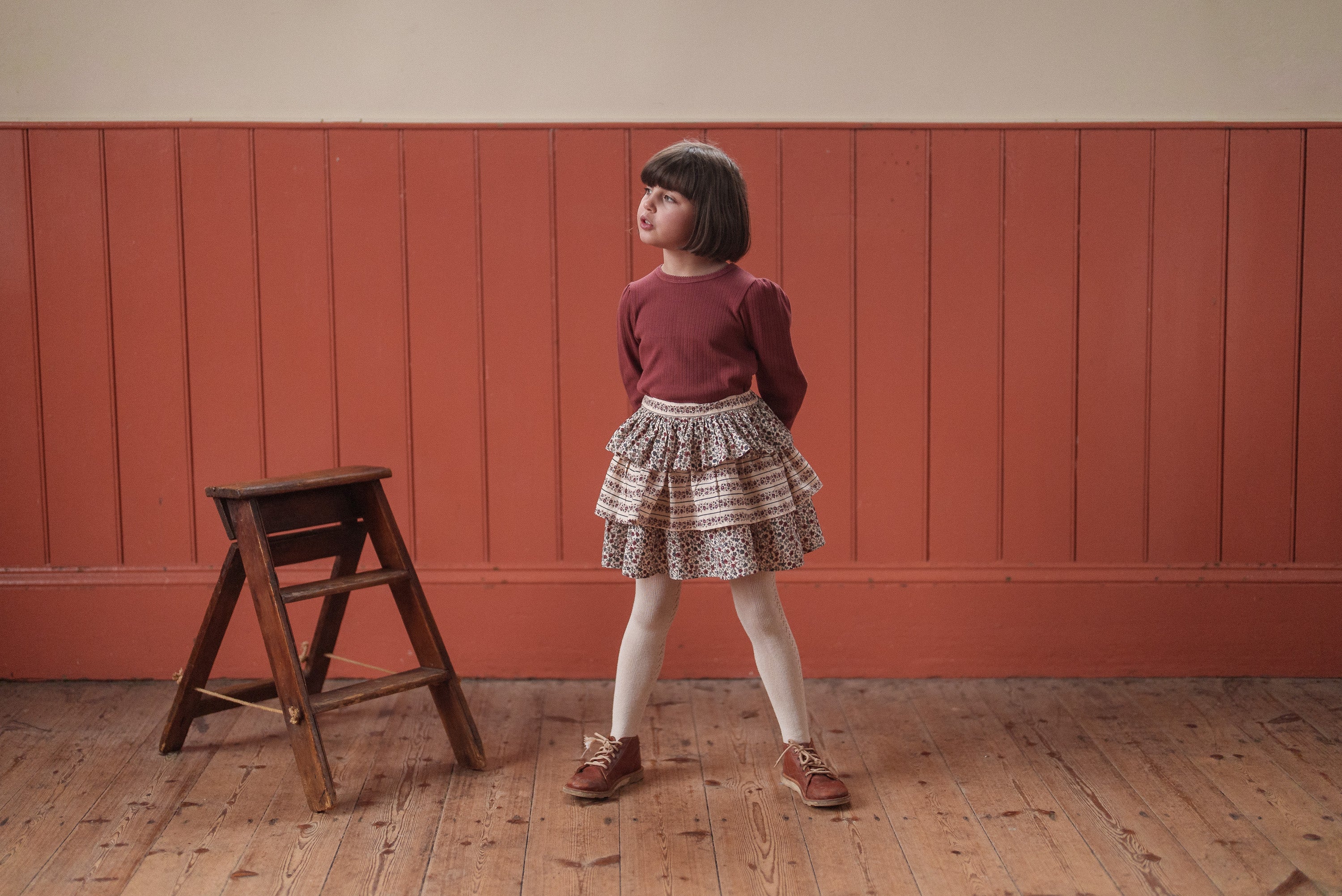 Young girl wearing a red organic cotton T-shirt with a vintage-style layered skirt, standing on a wooden floor next to a small wooden stool in a cozy, natural setting.
