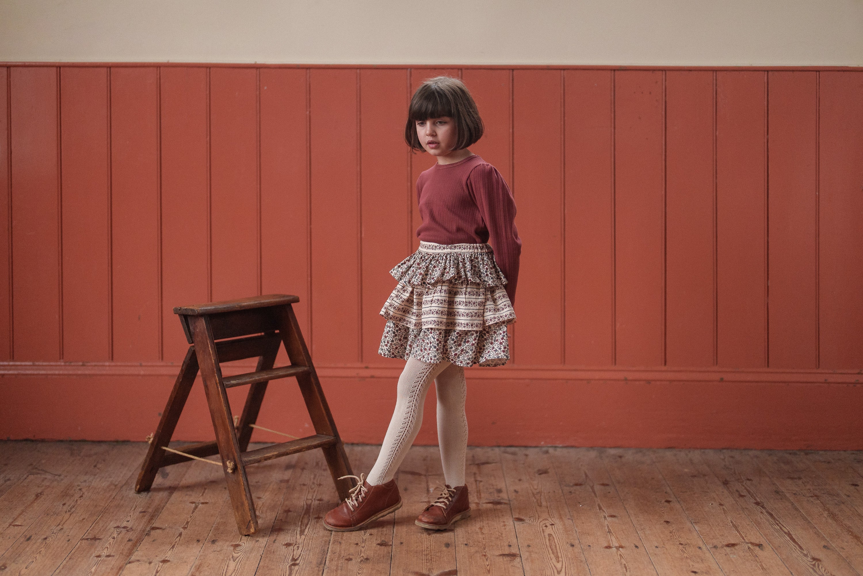 Young girl wearing a red organic cotton pointelle top and vintage-style layered skirt, standing on wooden floor beside a small wooden stool, showcasing sustainable kids clothing.