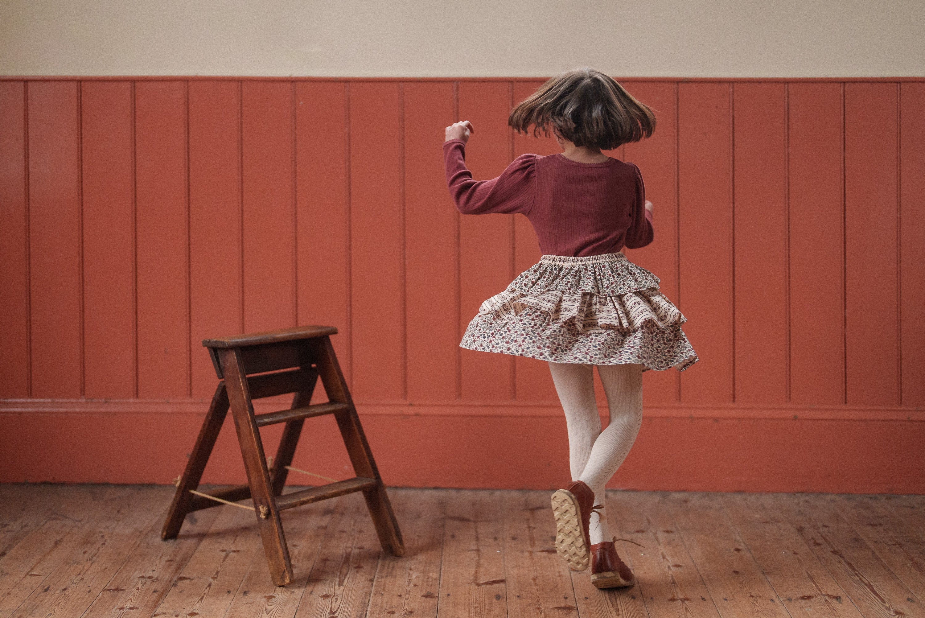 Young child wearing a red organic cotton top and vintage-style ruffled skirt, playfully jumping inside a cozy, natural wood-floored room with warm red paneled walls.