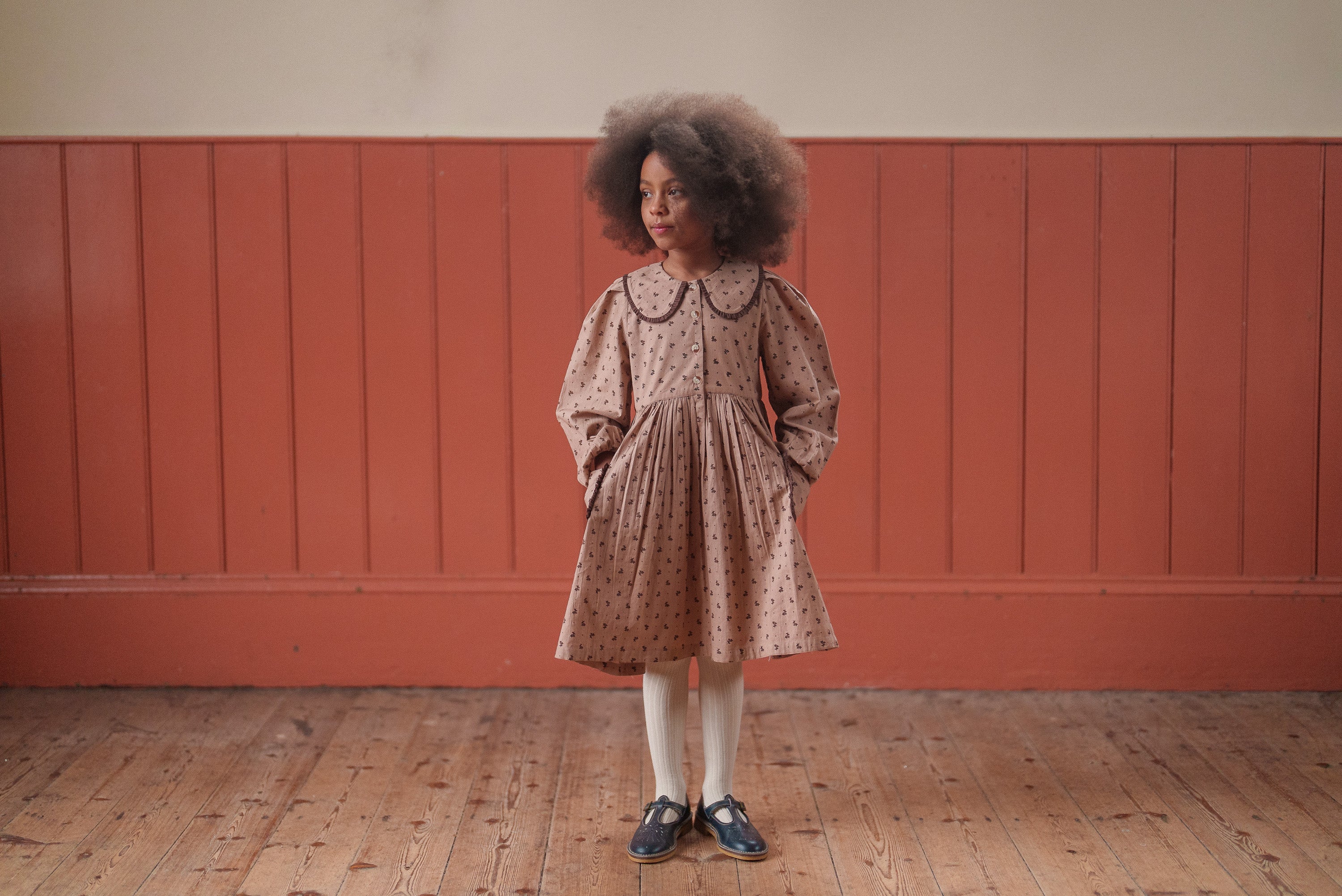 Young girl wearing a sustainable vintage-style dress in organic cotton with a subtle floral pattern, standing in a softly lit room with wooden floors and walls.