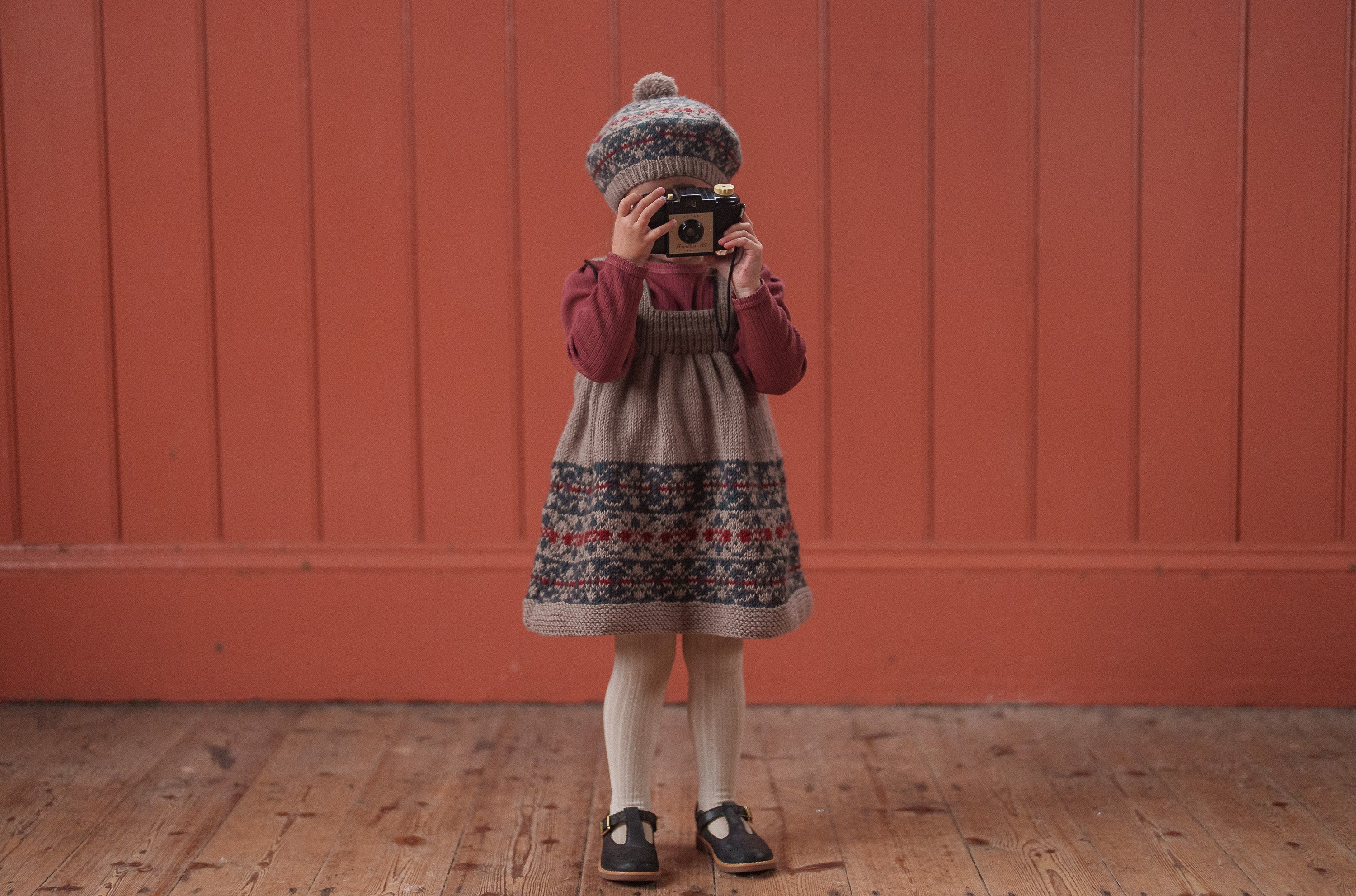 Child wearing vintage style knitted dress and patterned hat, holding a camera against a red natural wooden wall, showcasing sustainable children's clothing.