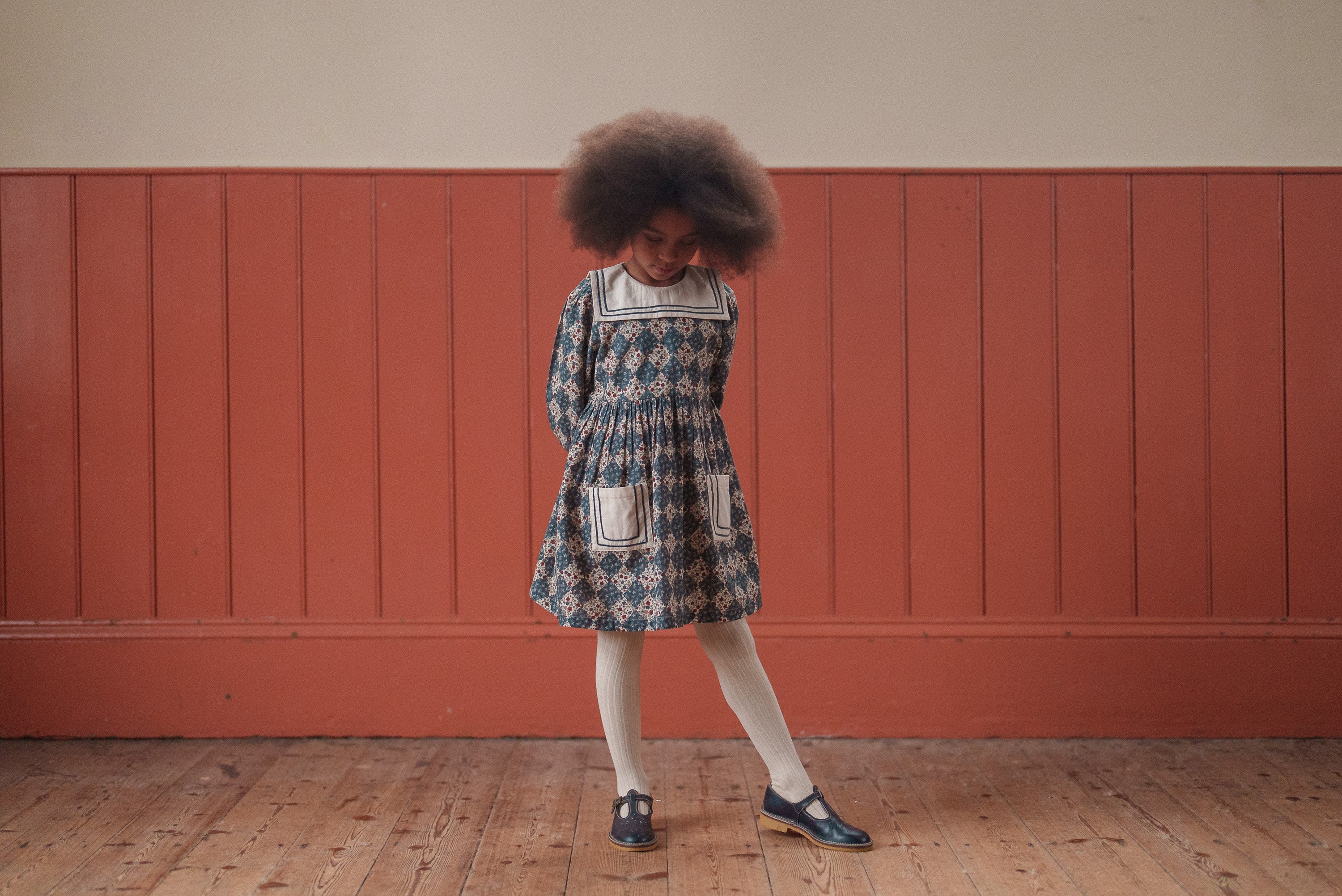 Young girl wearing a navy floral organic cotton dress with a vintage style, standing on a wooden floor against a warm-toned paneled wall, showcasing sustainable children's clothing.
