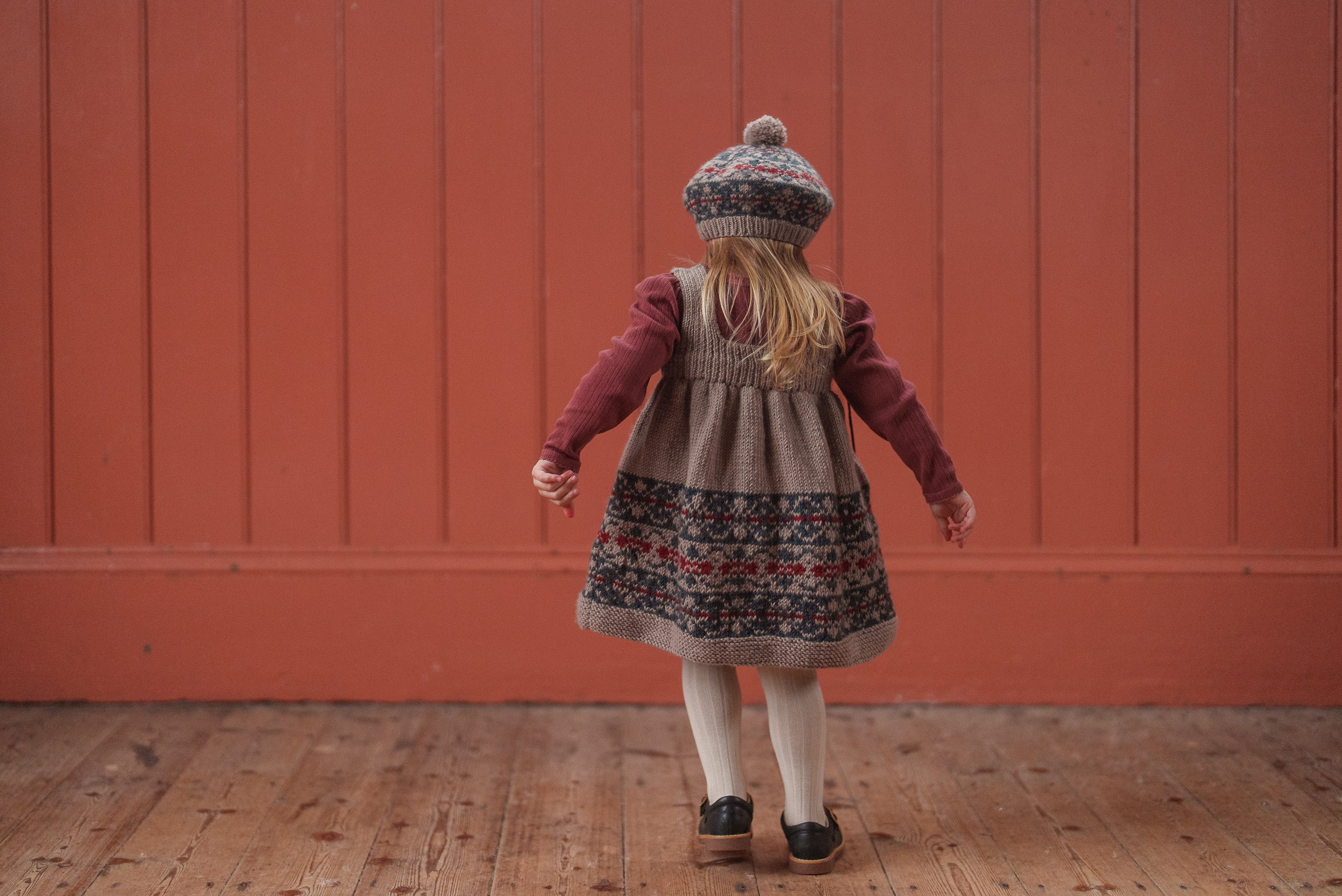 Child wearing vintage style dress and multicolor hand-knitted Merino beret, standing on wooden floor against red wall, showcasing natural and sustainable kids clothing.