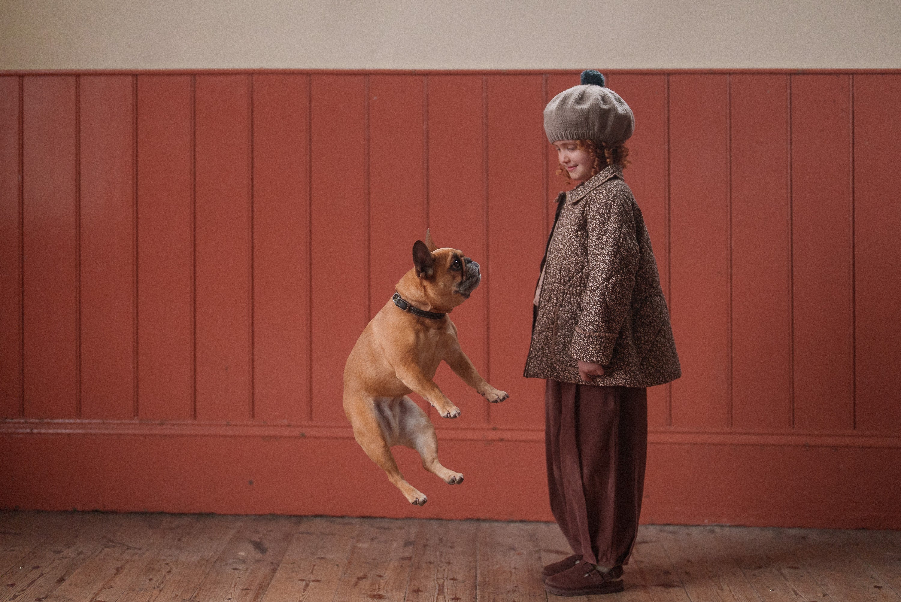 A child wearing vintage-style, ethically made clothing and a cozy hand-knitted beret in soft merino wool, happily interacting with a playful dog indoors.