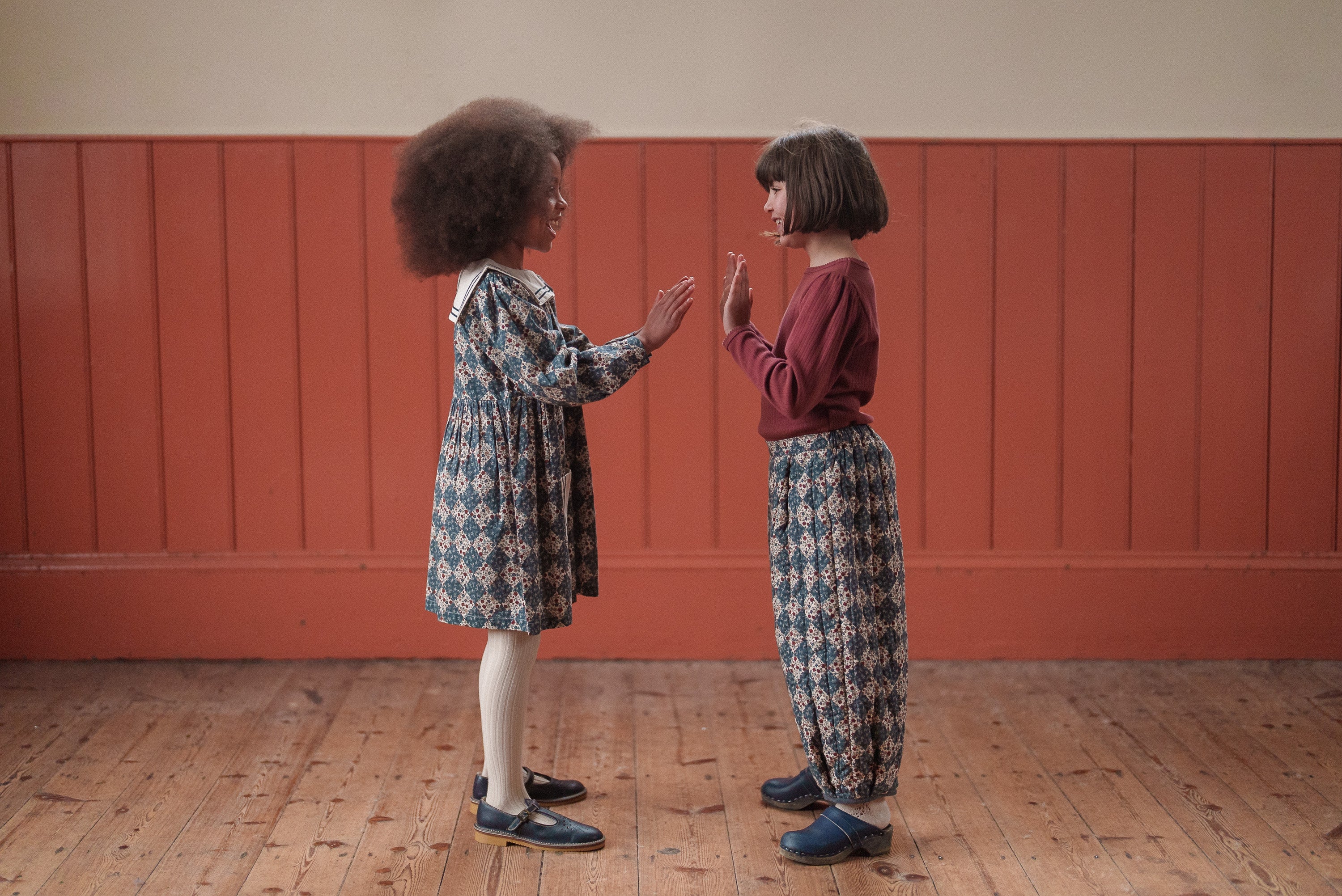 Two young girls facing each other indoors, wearing vintage style sustainable children’s clothing made from organic cotton in a warm, softly lit room with wooden floors and red paneling.