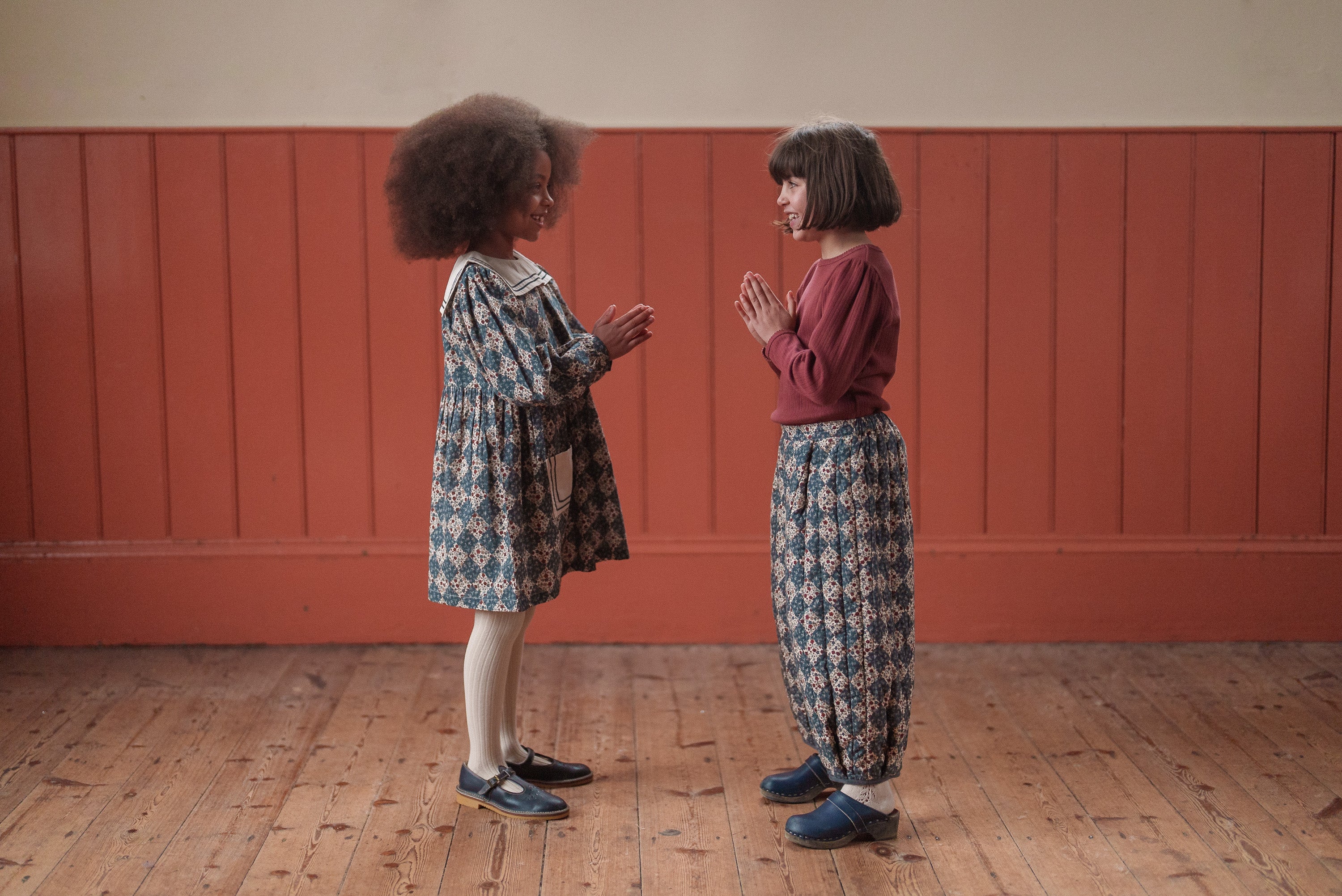 Two young girls facing each other indoors wearing vintage style children's clothing made from organic cotton with floral navy patterns, reflecting natural and ethical kidswear.