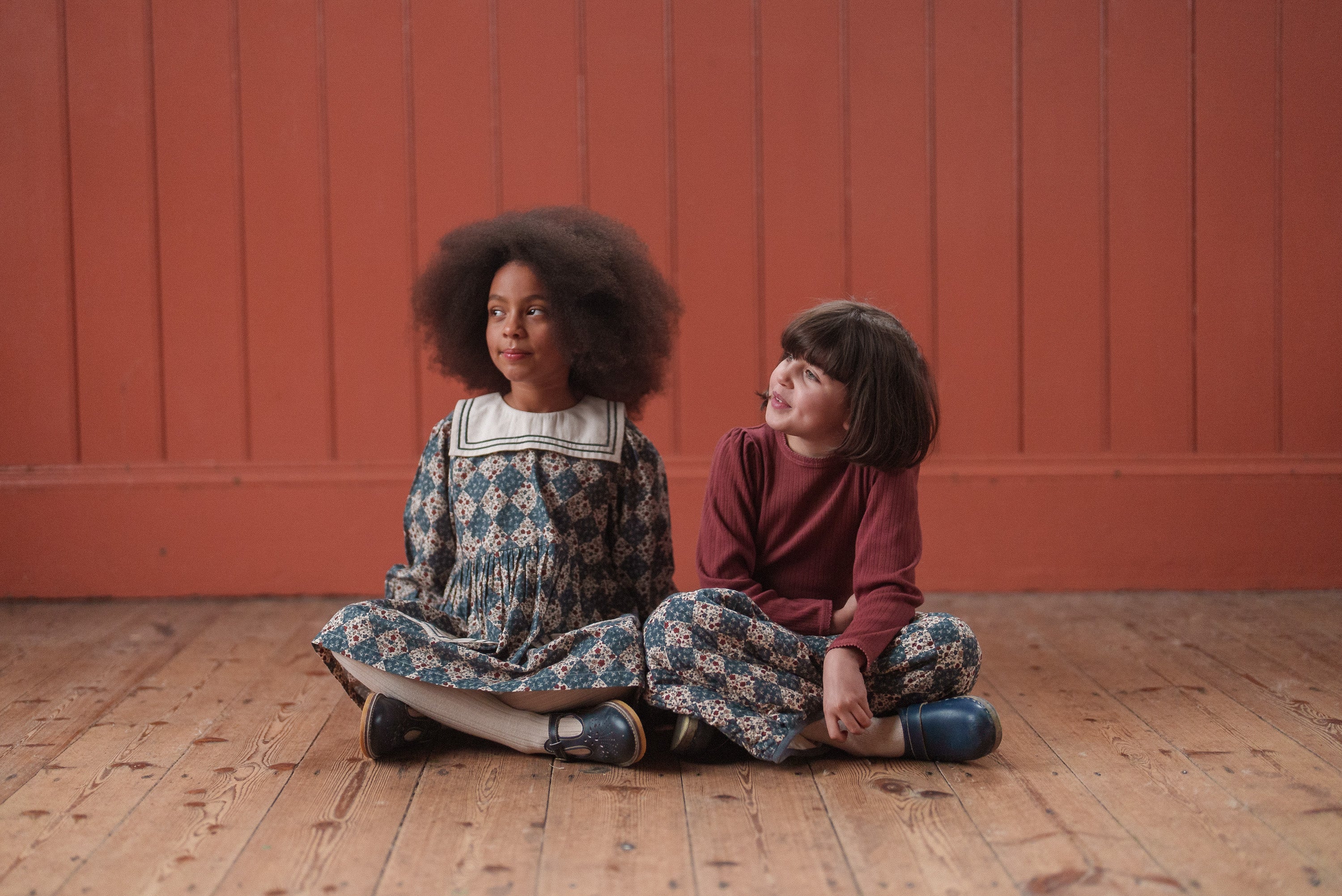 Two young girls sitting on a wooden floor against a warm orange wall, wearing vintage style, organic cotton children's clothing in navy and floral patterns, showcasing slow, natural and ethical fashion.