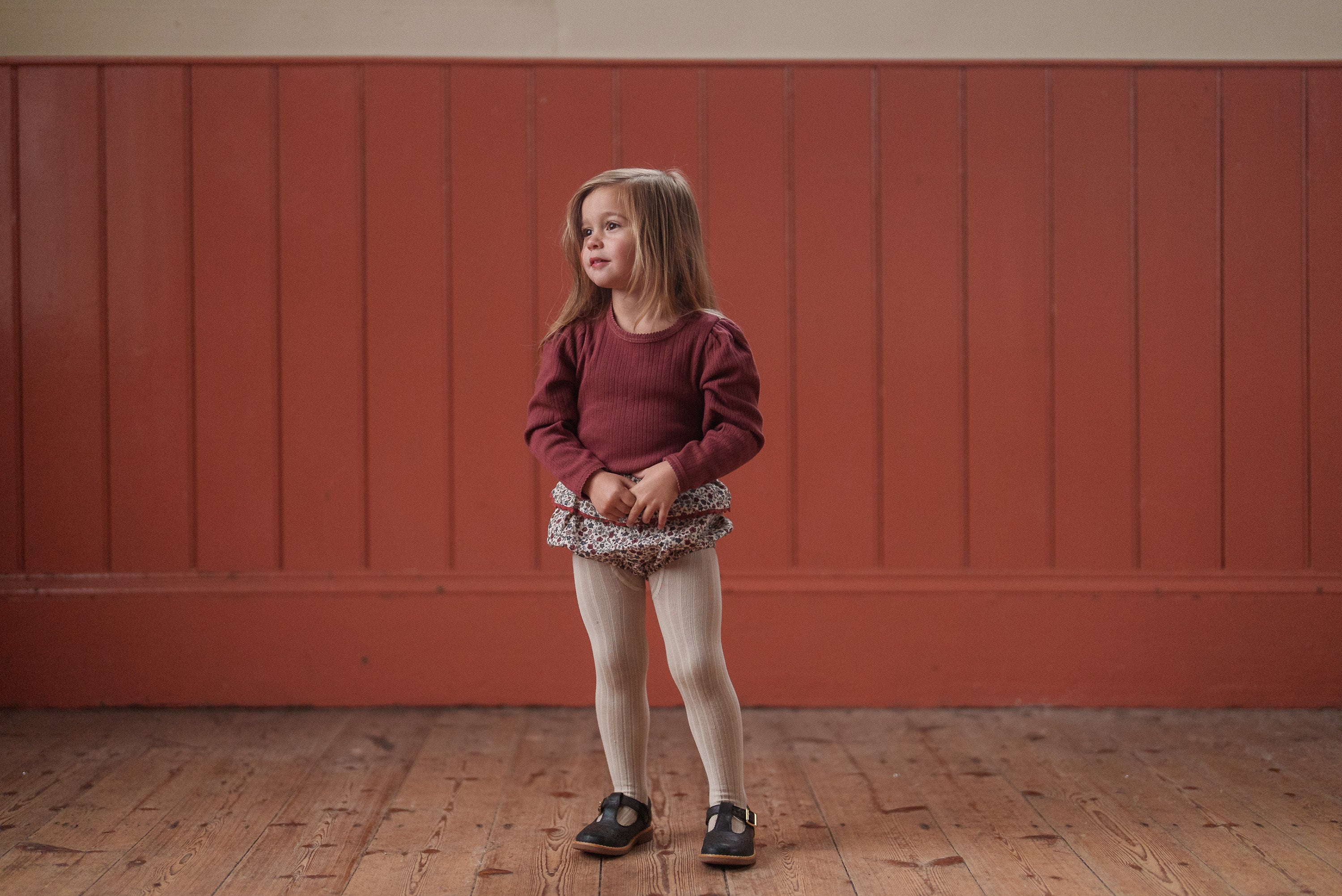 Young girl wearing a red organic cotton pointelle T-shirt paired with vintage-style floral shorts and cream tights in a warm, natural setting.