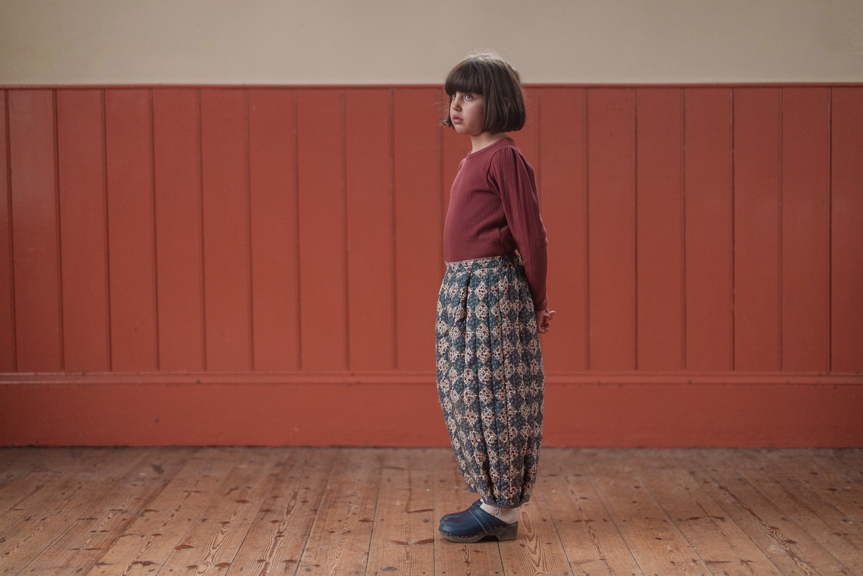 Child wearing vintage style quilted floral trousers made from organic natural materials, standing against a warm wooden panel wall in a cozy room.