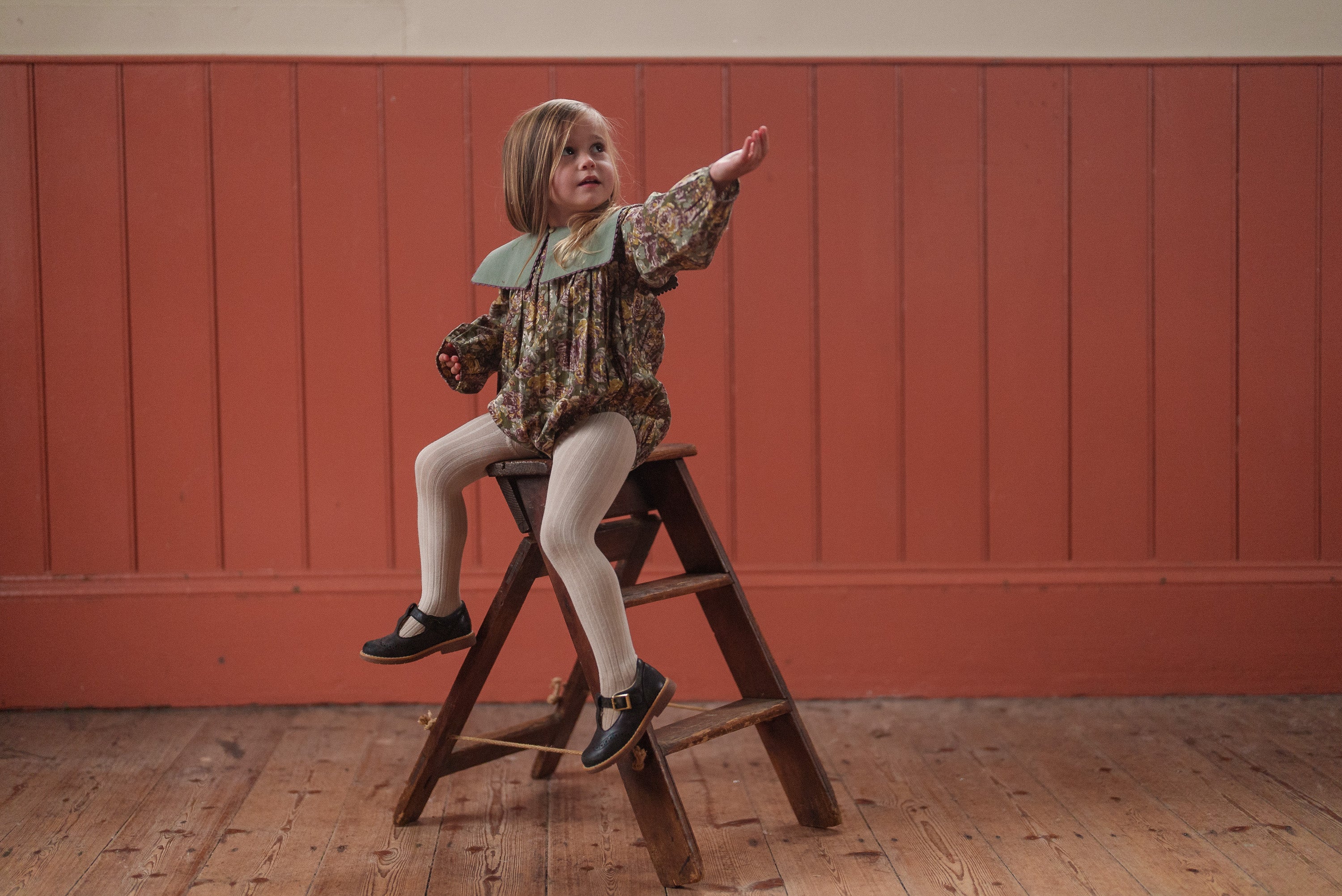 Young child wearing a vintage style floral romper in green and pink organic cotton, sitting on a wooden stool against a warm orange paneled wall, showcasing sustainable kids clothing.