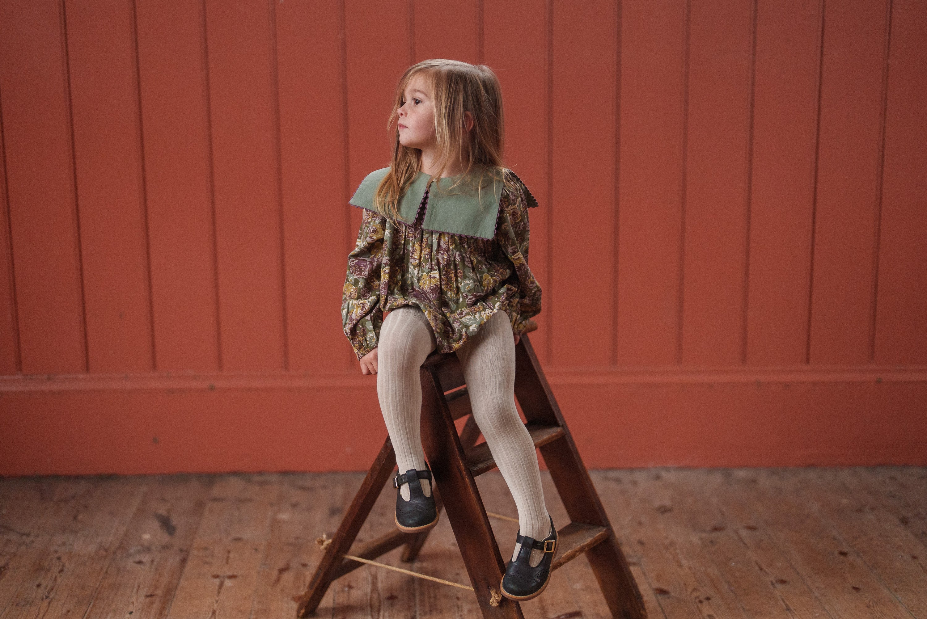 Young girl wearing a vintage style organic cotton romper with floral and green details, sitting on a wooden stool against a warm orange paneled wall in sustainable kids clothing.