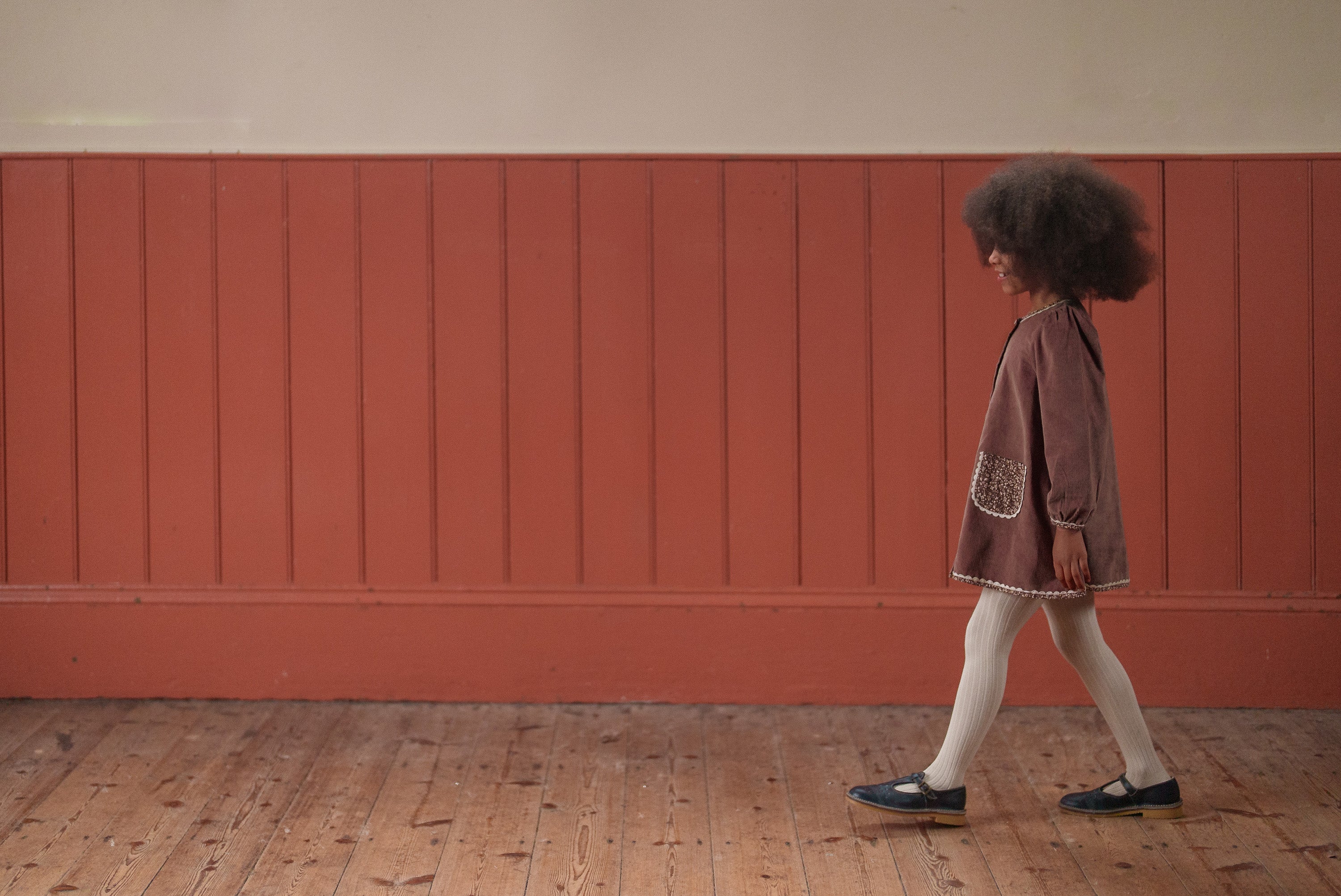 Young girl walking indoors wearing a vintage-style nutmeg velvet dress with floral pockets, paired with white tights and black shoes, made from organic cotton.
