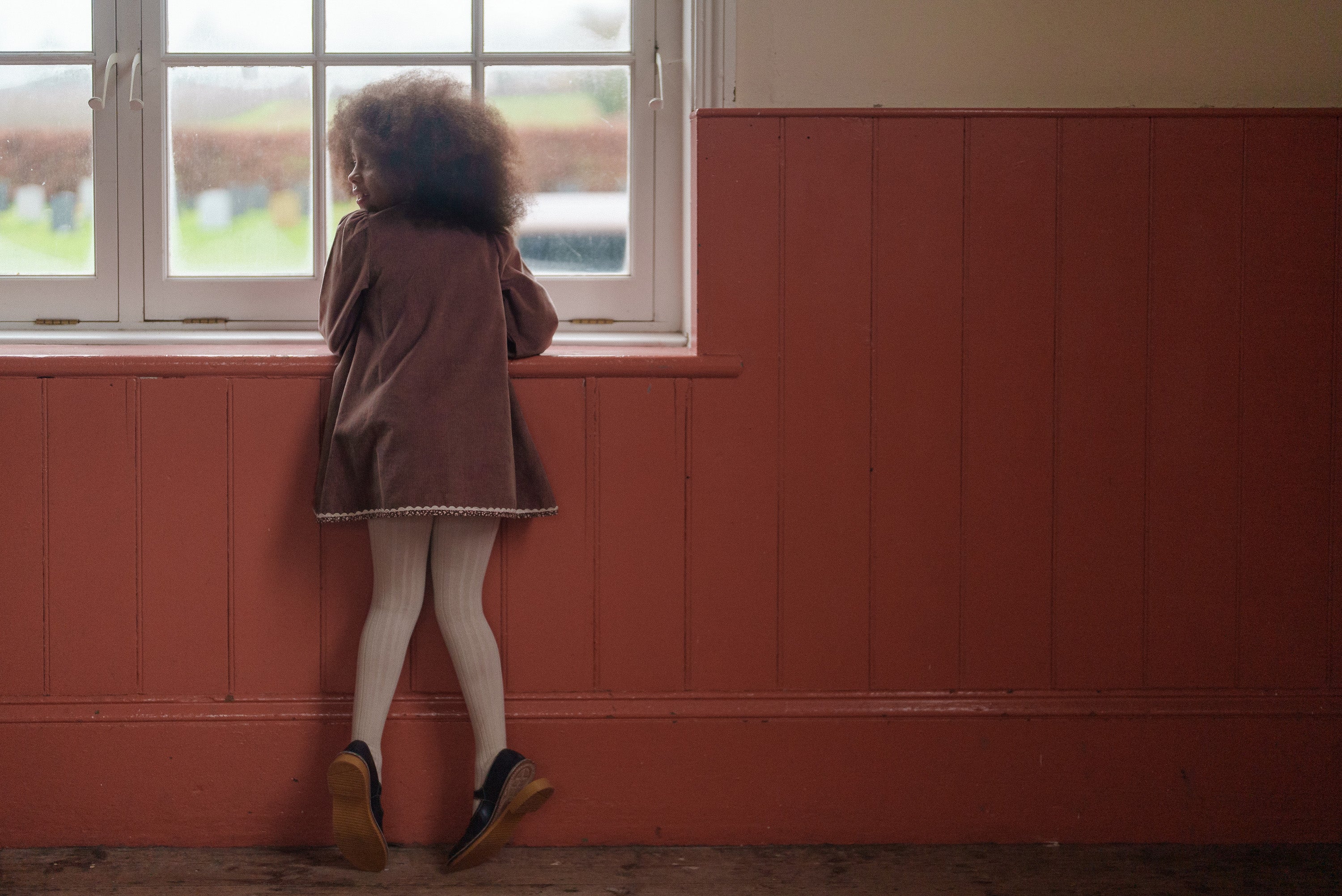 Child wearing a vintage style nutmeg velvet dress with floral details made from organic cotton, standing by a window in a cozy room with natural light.