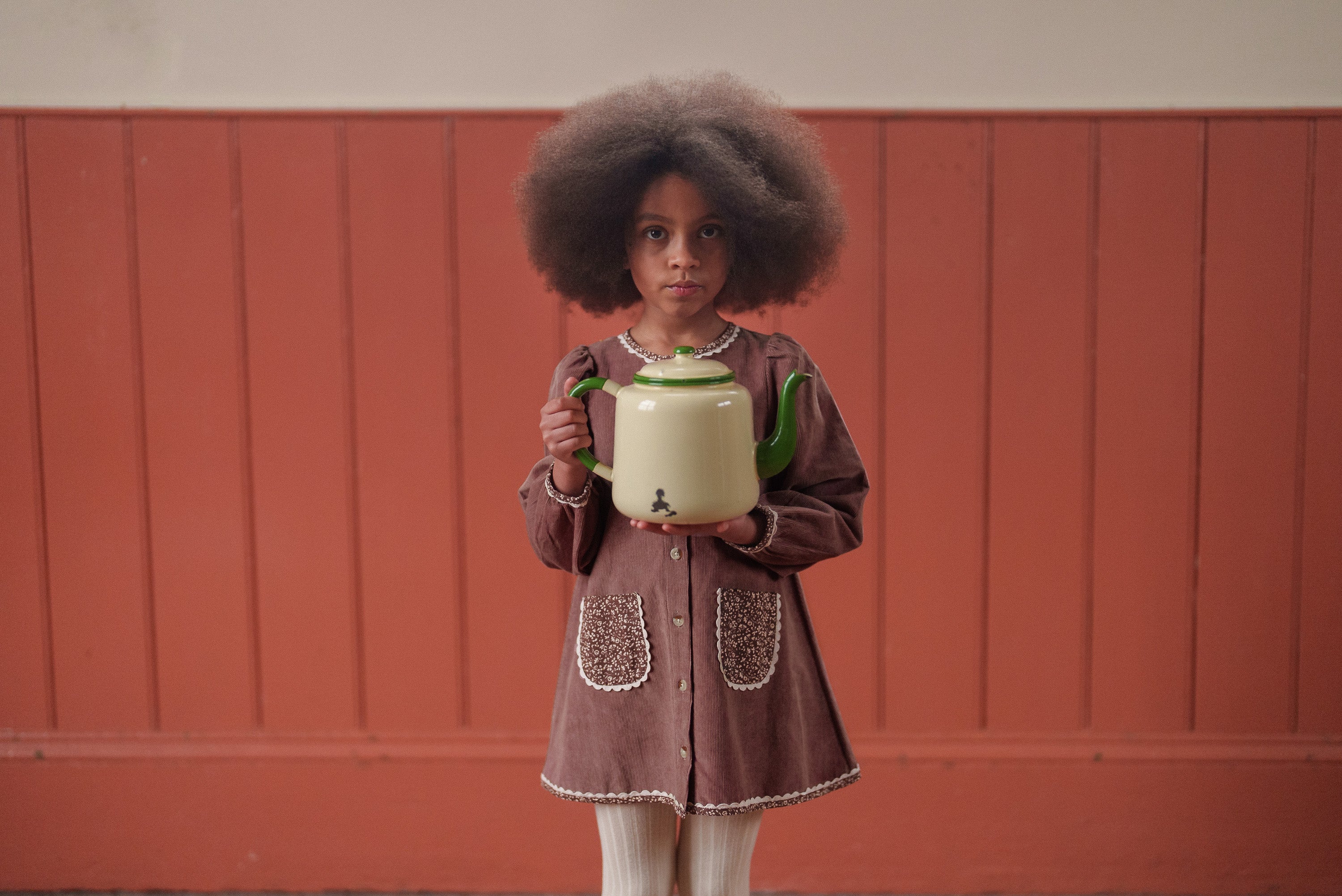 Young girl with natural hair wearing a vintage style organic cotton velvet dress in nutmeg tone with floral accents, holding a green vintage kettle against a warm background.