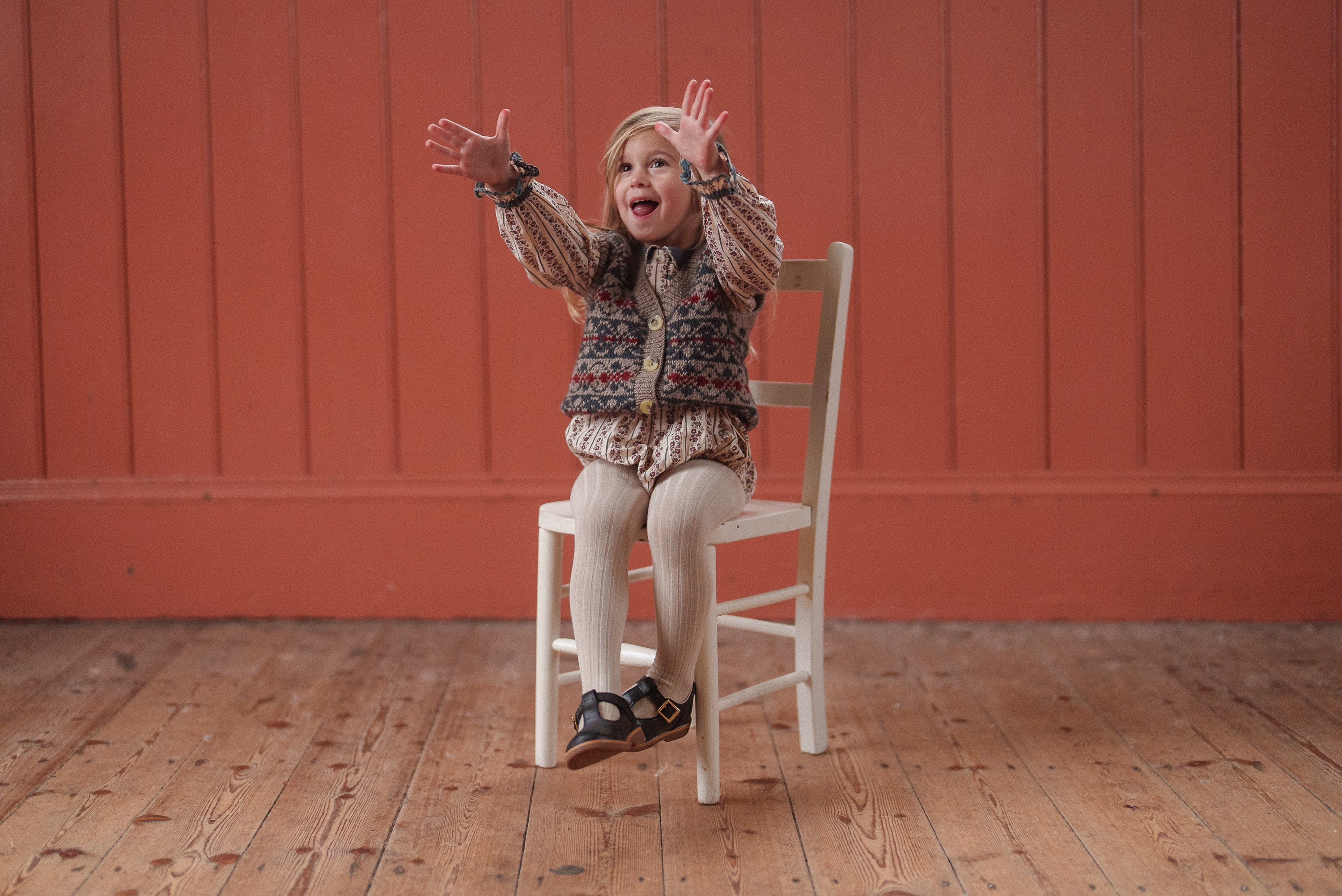 A joyful toddler girl wearing a vintage style romper and knitted vest made from organic cotton, sitting on a wooden chair against a warm orange wooden wall.