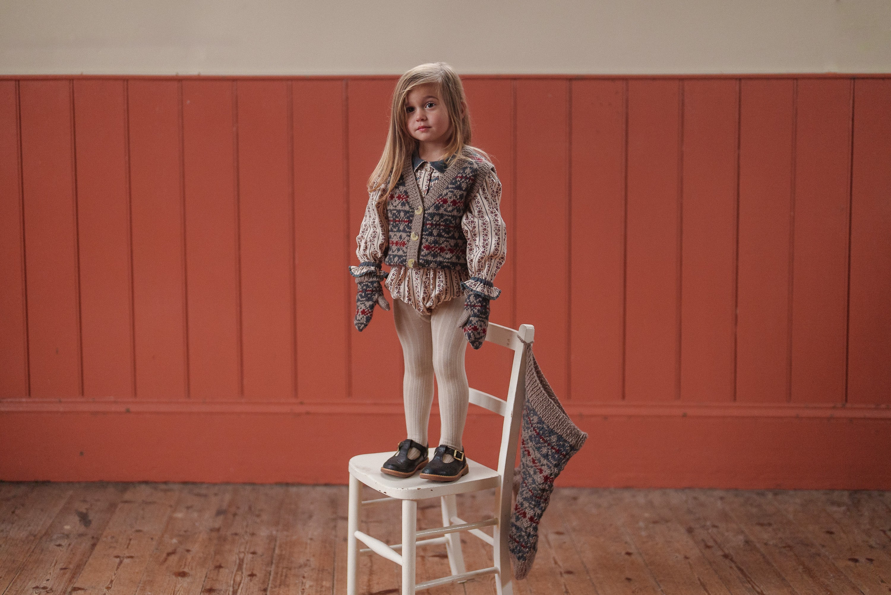 Young girl wearing a vintage-style organic cotton romper with floral patterns, standing on a white wooden chair in a warm, natural-toned room.