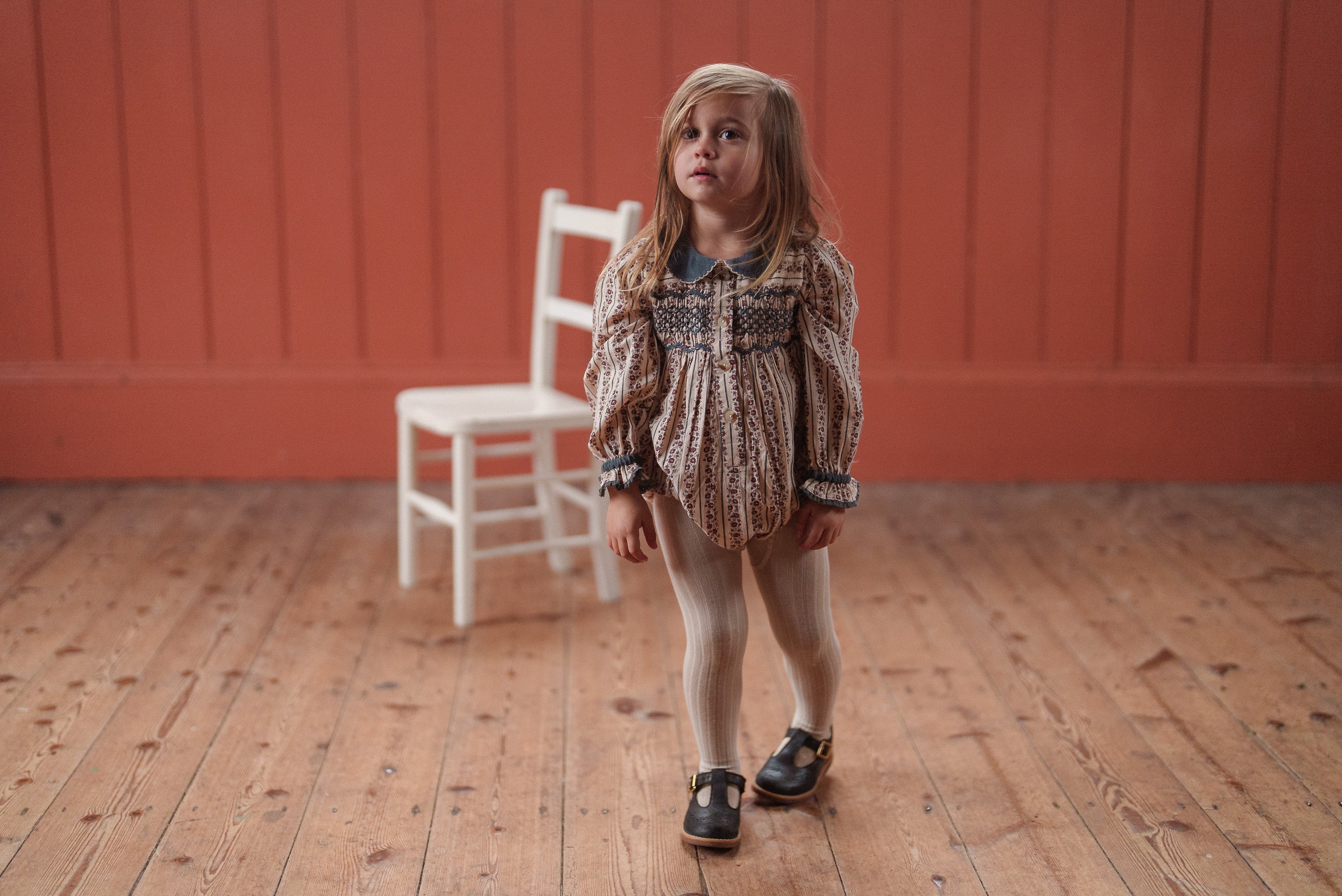 Young girl wearing a vintage-style romper made from organic cotton in a soft floral pattern, standing on a wooden floor with a white chair in the background.