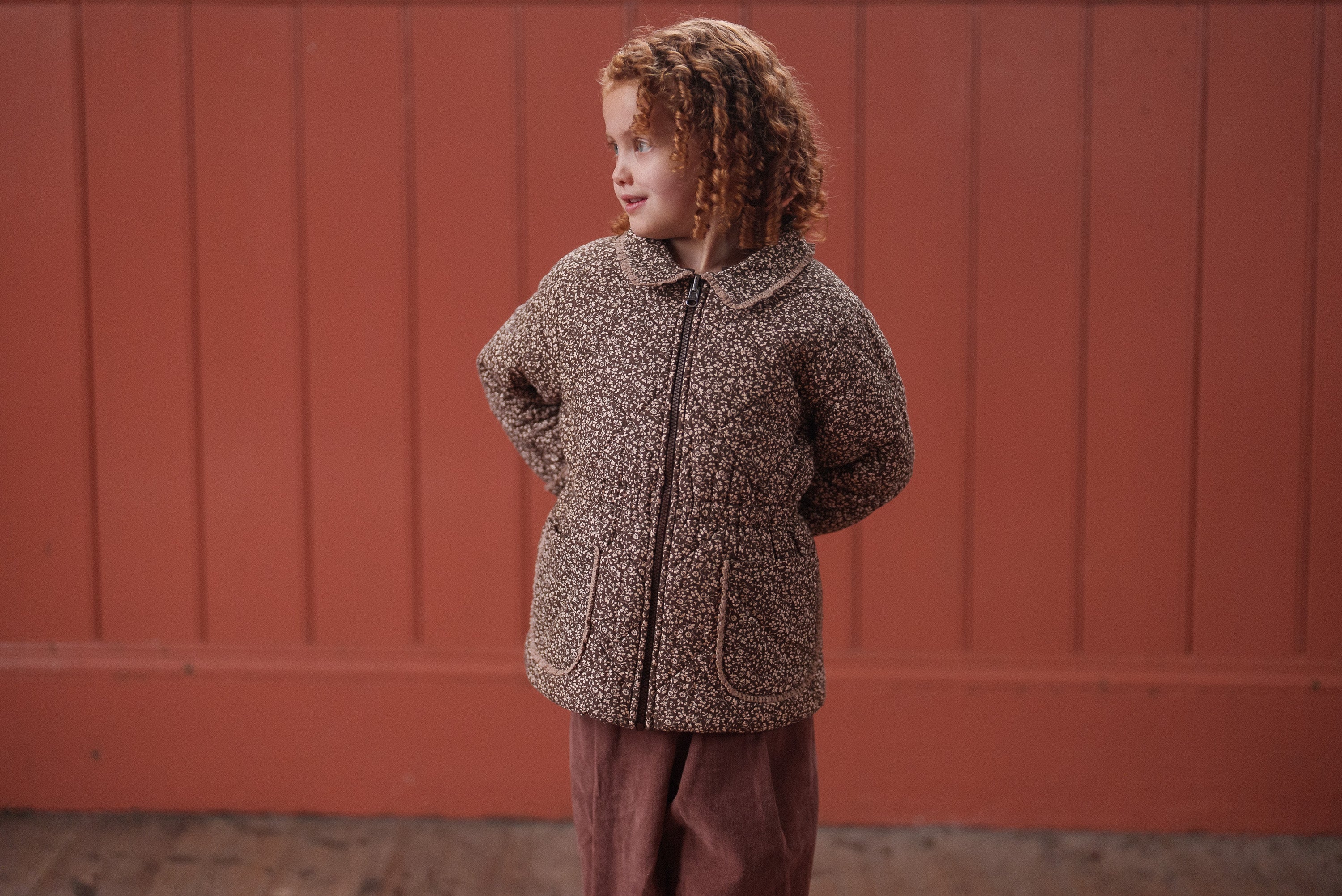 Child wearing a brown and beige floral organic cotton jacket with pockets, styled in a vintage-inspired, naturally made kids jacket against a warm-toned wooden background.