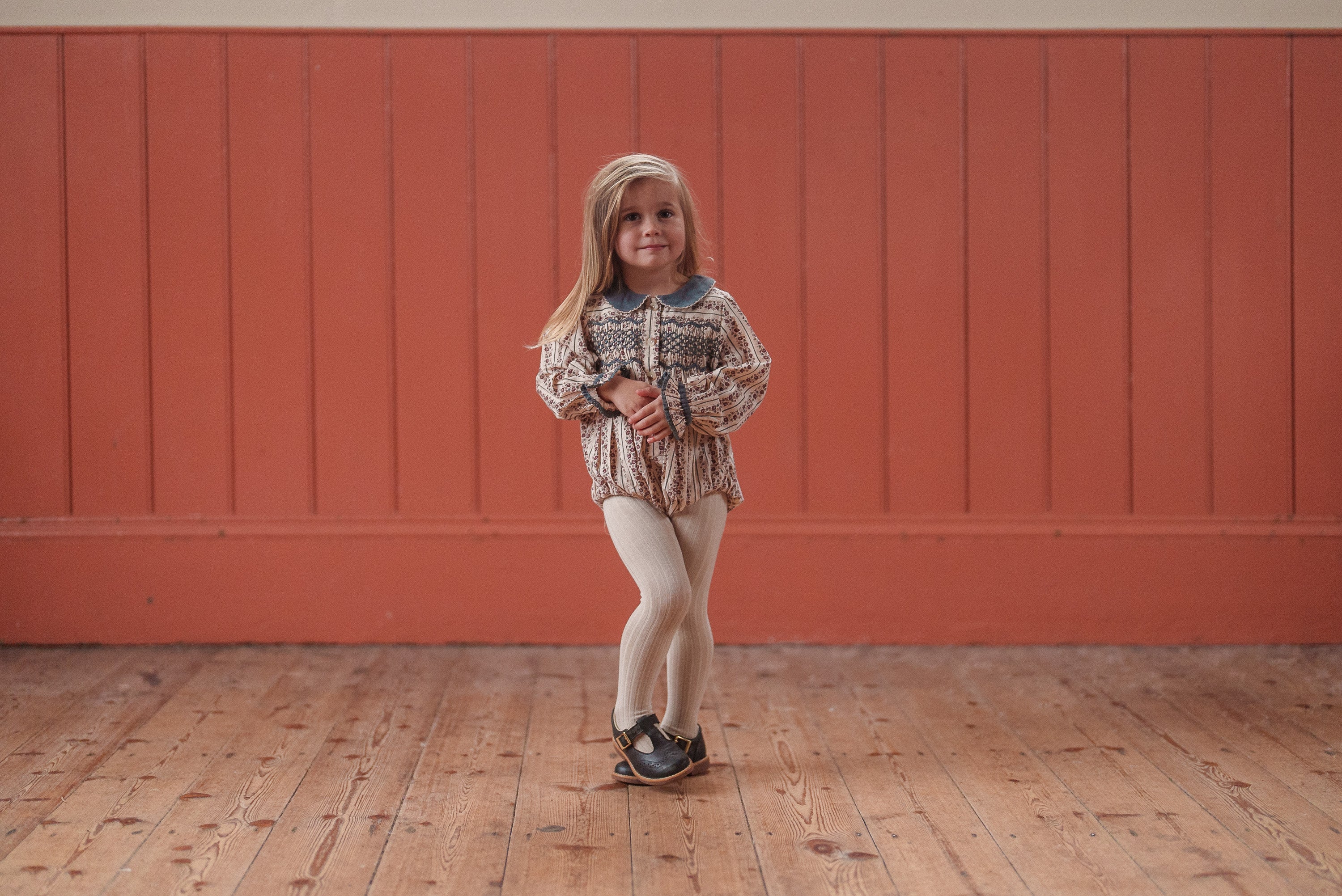 Young girl wearing a natural, vintage-style romper made from organic cotton, standing on a wooden floor with a warm, rustic background, embodying ethical, sustainable kids clothing.