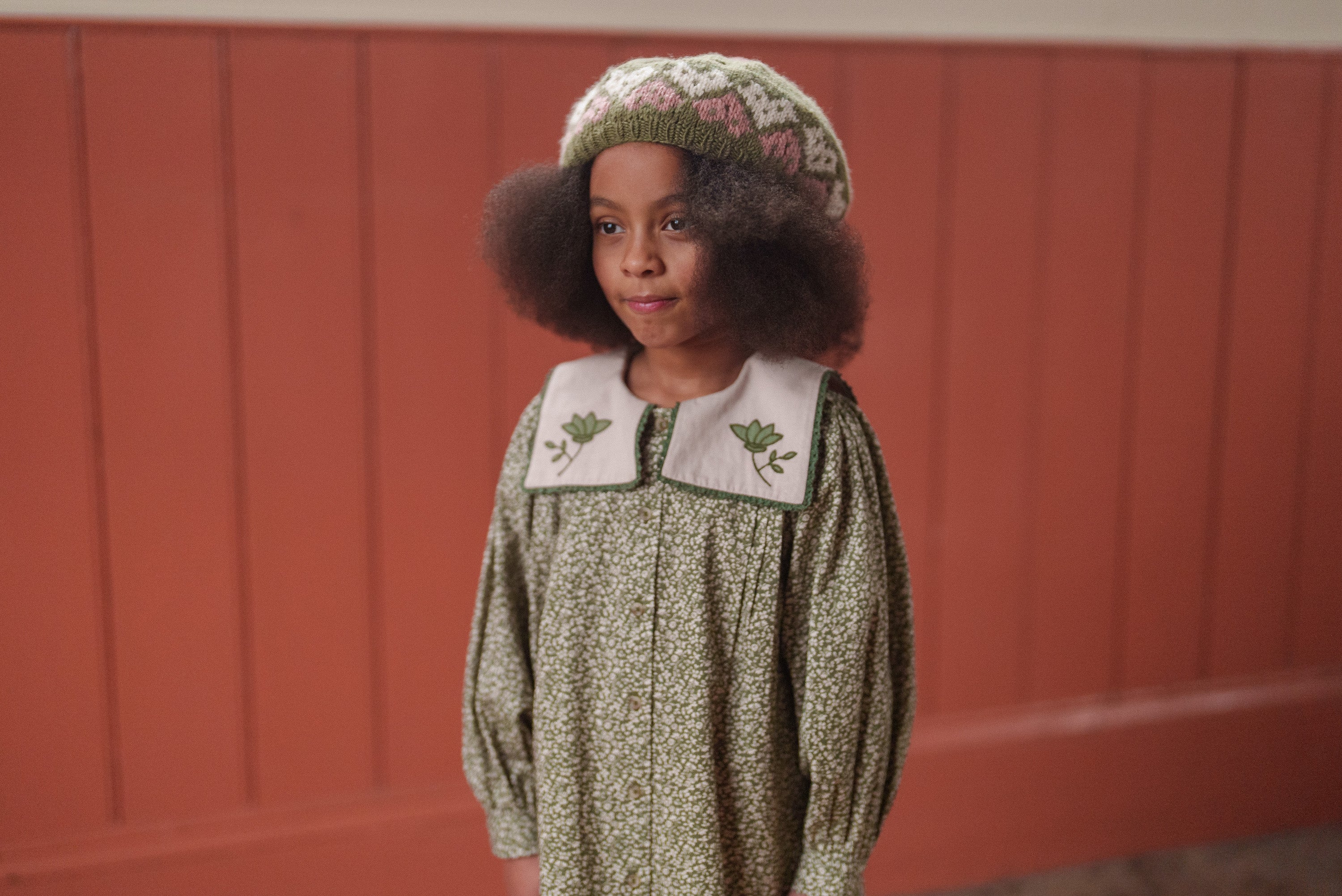 Young girl wearing a vintage style green floral organic cotton dress with a detailed collar and a knitted hat, reflecting sustainable and ethically made children’s clothing.